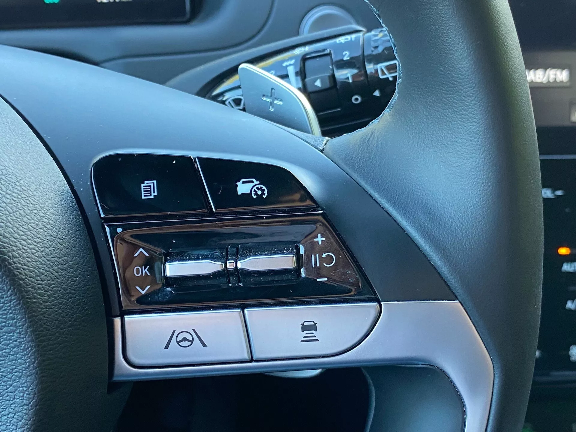 Blízky pohľad na ovládacie prvky na volante na ľavej strane Hyundai Tucson 1.6 T-GDI Pure z roku 2020, ukazujúce funkcie ako navigácia v menu, ovládanie hlasitosti, aktivácia hlasových príkazov a funkcia Bluetooth pre telefonovanie.