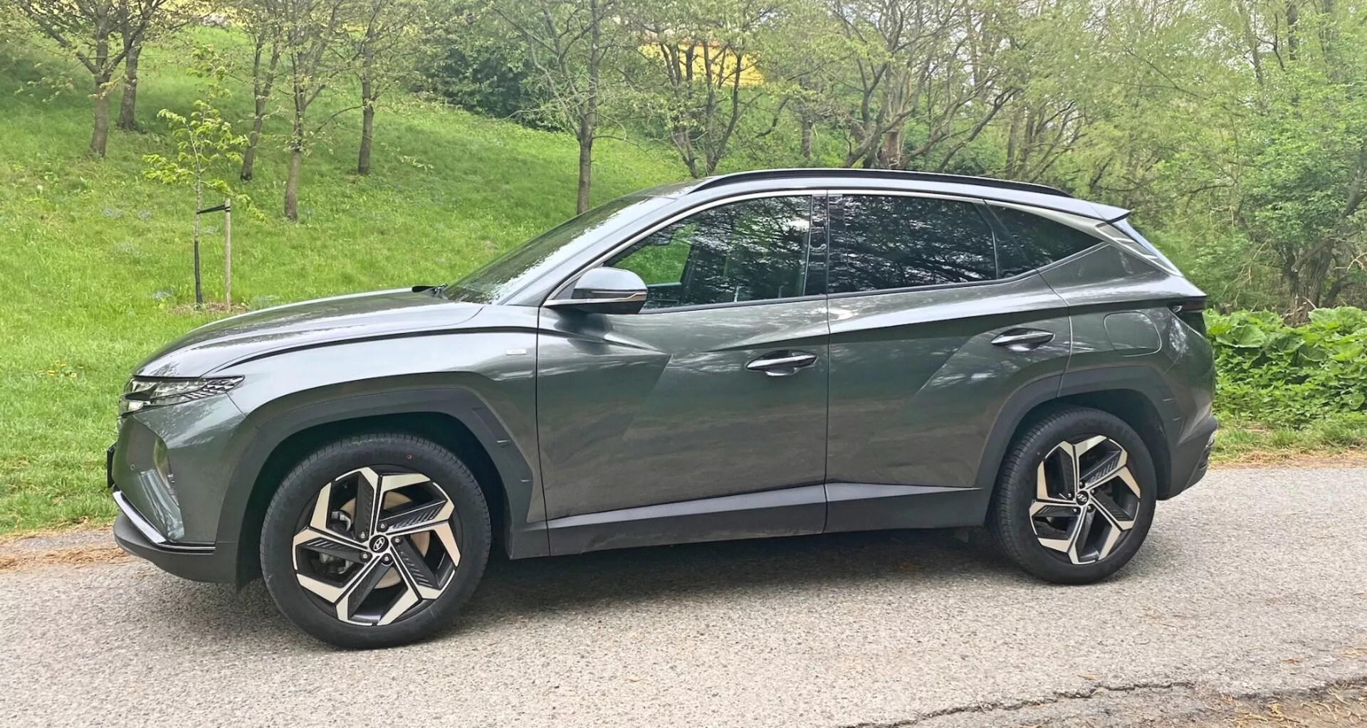 Bočný profil moderného crossover SUV Hyundai Tucson 2020 1.6 T-GDI Pure v metalickej šedej farbe s lesklým povrchom. Vozidlo má veľké, zložité viacramenné zliatinové kolesá, LED svetlomety, mierne tónované okná, aerodynamické línie karosérie so sklonom strechy končiacej zadným spoilom, strešné nosiče, bočné zrkadlá s integrovanými smerovkami a chrómové alebo kovové akcenty. Vozidlo je umiestnené na asfaltovej ceste s miernym sklonom nadol a umiestnené skôr vpravo od rámu obrazu.