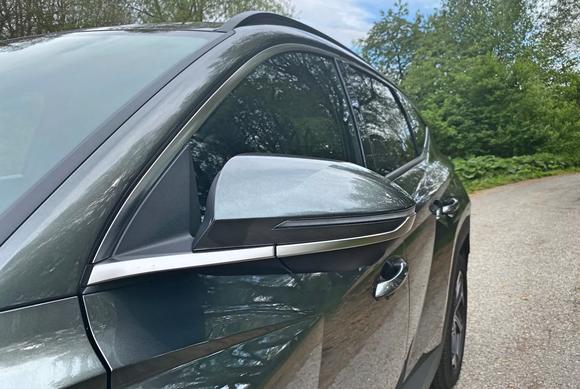 Detailný bočný pohľad na Hyundai Tucson 2020 1.6 T-GDI Pure zvýrazňuje jeho moderný dizajn a technologické prvky, so zameraním na stranu vodiča. Fotografia z nízkeho uhla zdôrazňuje elegantné, aerodynamické spätné zrkadlo s integrovanými smerovými ukazovateľmi a čiernymi prvkami, ktoré naznačujú pokročilé funkcie. Zapustená predná kľučka dverí vodiča naznačuje bezkľúčový vstup, čím prispieva k aerodynamickej účinnosti vozidla. Lesklé, telo farbené dvere a okenné lišty s chrómom naznačujú prémiovú výbavu. Prvky pozadia, ako sú stromy a obloha, naznačujú vonkajšie prostredie.