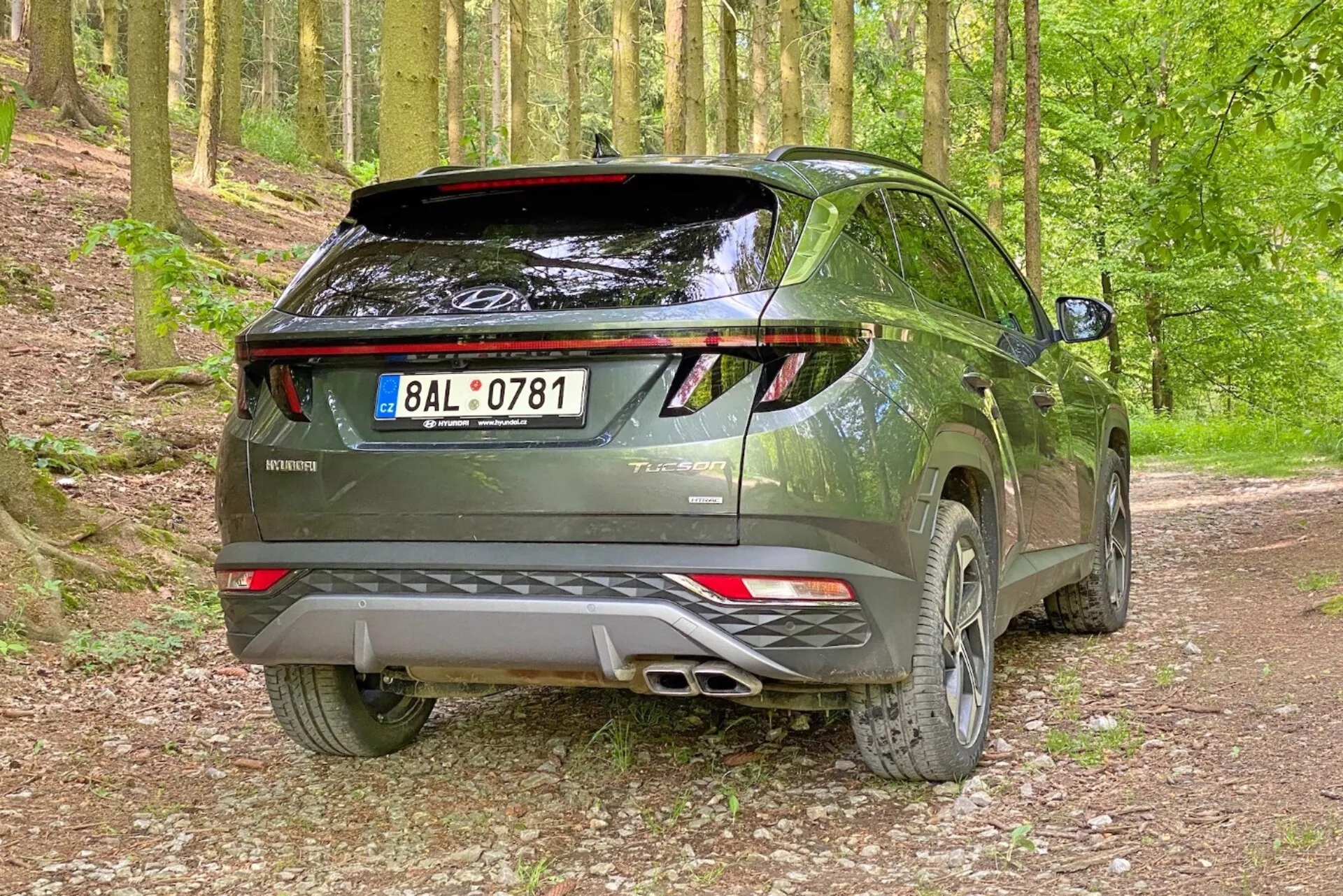 Obrázok moderného SUV Hyundai Tucson 2020 v lesnom prostredí na štrkovej ceste, zachytený z pravého zadného trojštvrťového uhla. Fotografia zvýrazňuje pravú stranu a zadný profil vozidla s viditeľnými zadnými dverami, LED zadnými svetlami, dvojitým výfukovým systémom, zadným spojlerom a zliatinovými kolesami.