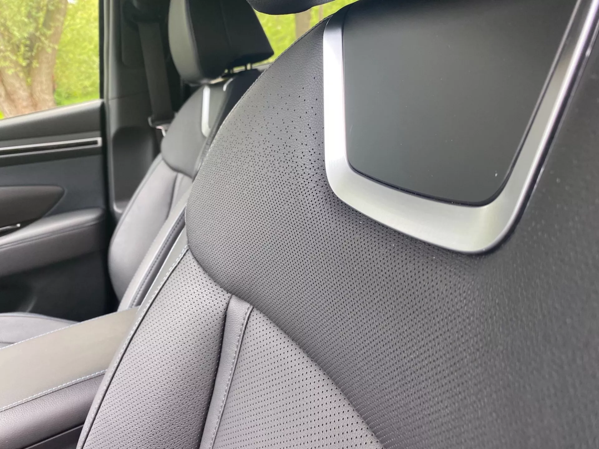 Detailný pohľad na interiér Hyundai Tucson 2020, zameraný na pravý A-stĺpik a časť pravého predného sedadla. A-stĺpik je pokrytý textúrovaným materiálom, pravdepodobne látkou alebo syntetickou kožou, čo naznačuje estetické alebo hluk redukčné zameranie. Sedadlo má dvojfarebný dizajn a je perforované pre ventiláciu. Výrazným prvkom na križovatke A-stĺpika a stropnej výplne môže byť súčasťou audio systému, komponentom airbagu alebo senzorom asistencie vodiča, zdôrazňujúcim pokročilé bezpečnostné alebo technologické funkcie.
