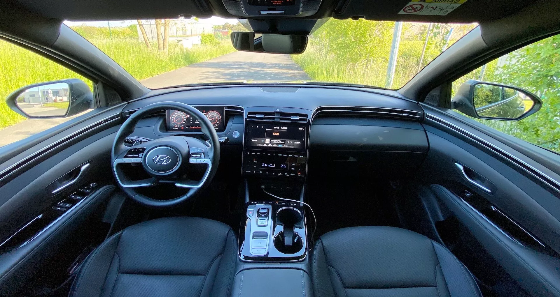 Interiér Hyundai Tucson 2020 z pohľadu zadného sedadla s pohľadom na prednú prístrojovú dosku, volant s ovládacími prvkami, digitálny prístrojový panel, veľký dotykový informačný systém, tlačidlá na ovládanie klimatizácie, radiacu páku a predné sedadlá potiahnuté kožou za prirodzeného denného svetla.