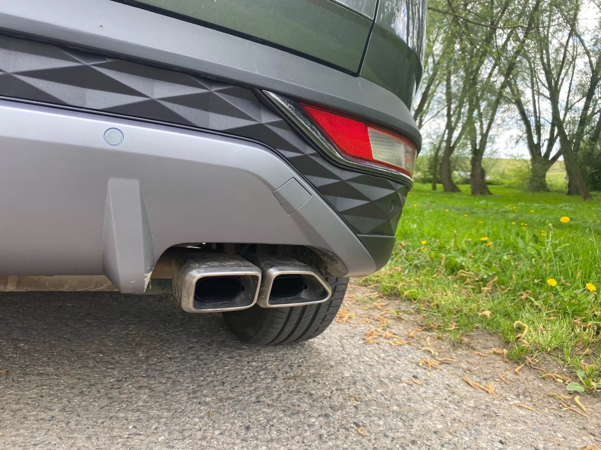 Detailný záber zadného rohu vozidla Hyundai Tucson 2020 z úrovne zeme. Viditeľné detaily zahŕňajú dvojfarebný zadný nárazník, čiastočné LED zadné svetlo a kovové dvojité výfukové potrubie s obdĺžnikovými koncovkami, čo naznačuje športovú výbavu. Elegantný dizajn vozidla a metalický lak odrážajú okolie.