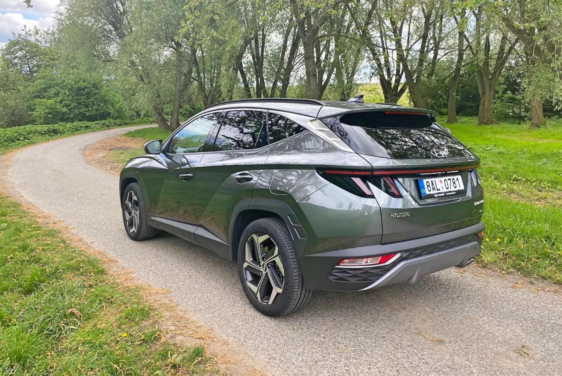 Obrázok zobrazuje moderné crossover SUV z uhla tri štvrtiny zozadu, kde je vidieť elegantný, aerodynamický dizajn a sklony strechy. Tmavozelené vozidlo sa vyznačuje zdvihacími zadnými dverami, športovým spojlerom a veľkými hliníkovými kolesami s viaceronásobným dizajnom lúčov. Viditeľné prvky zahŕňajú moderné zadné LED svetlá, dvojité výfukové potrubie a stierač zadného okna. SUV je zaparkované na štrkovej ceste obklopenej zeleňou, čo naznačuje park alebo vidiecke prostredie.