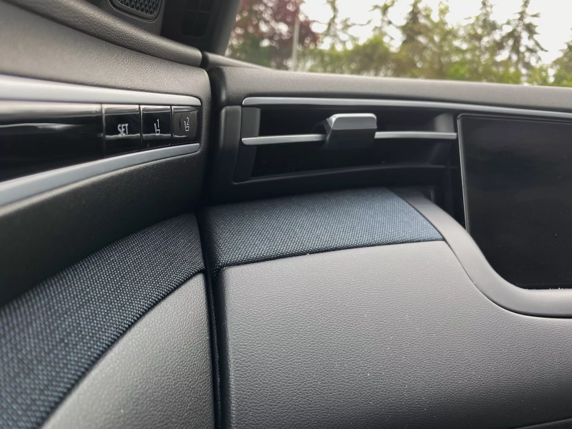 Detail interiéru Hyundai Tucson 2020, zobrazujúci tmavo sfarbenú, textúrovanú stranu palubnej dosky na strane spolujazdca a časť dverového panelu. Scéna zvýrazňuje niekoľko ovládacích tlačidiel, nastaviteľný ventilačný otvor s horizontálnymi lamelami, uzamykateľnú priehradku a zakrivený dizajn dverového panelu, ktorý plynule prechádza do palubnej dosky.