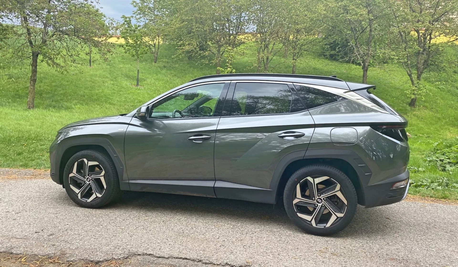 Bočný profil 2020 Hyundai Tucson 1.6 T-GDI Pure SUV v prirodzenom dennom svetle. Vozidlo sa nachádza na trávnatej ploche s stromami v pozadí, čo naznačuje vidiecke alebo prímestské prostredie. Moderné dizajnové prvky zahŕňajú LED svetlomety, integrované smerovky v bočných zrkadlách, tónované okná, aerodynamické hliníkové kolesá a klesajúcu strechu ako u kupé. Elegantná karoséria s charakterovými líniami a zapustenými kľukami dverí naznačujú pokročilý systém bezkľúčového vstupu, zatiaľ čo zadný spojler naznačuje aerodynamické vylepšenia. Auto je centrálne umiestnené v ráme a poskytuje čistý, nebránený pohľad zo strednej výšky.