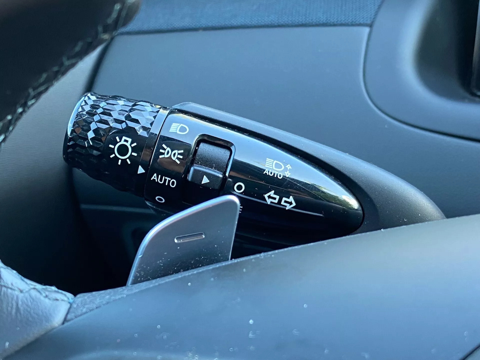 Detailný pohľad na ovládaciu páku umiestnenú na stĺpiku riadenia naľavo od volantu v Hyundai Tucson 2020. Páka obsahuje symboly na nastavenie svetlometov vrátane vypnutého stavu, automatických svetlometov, parkovacích svetiel a stretávacích/diaľkových svetiel. Obrázok zachytáva páku, časť volantu a rozmazané pozadie dverí vodiča, čím zdôrazňuje detail a kvalitu dizajnu. Polohu 'AUTO' indikuje automatický systém svetlometov.