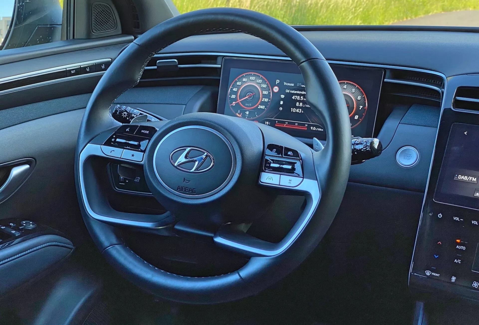 Interiér vozidla Hyundai Tucson 1.6 T-GDI 2020 z pohľadu vodiča so zameraním na moderný trojramenný volant s logom Hyundai, tlačidlá pre médiá a tempomat, čiastočne viditeľný prístrojový panel s tachometrom a otáčkomerom a centrálny dotykový infotainment systém na palubnej doske.
