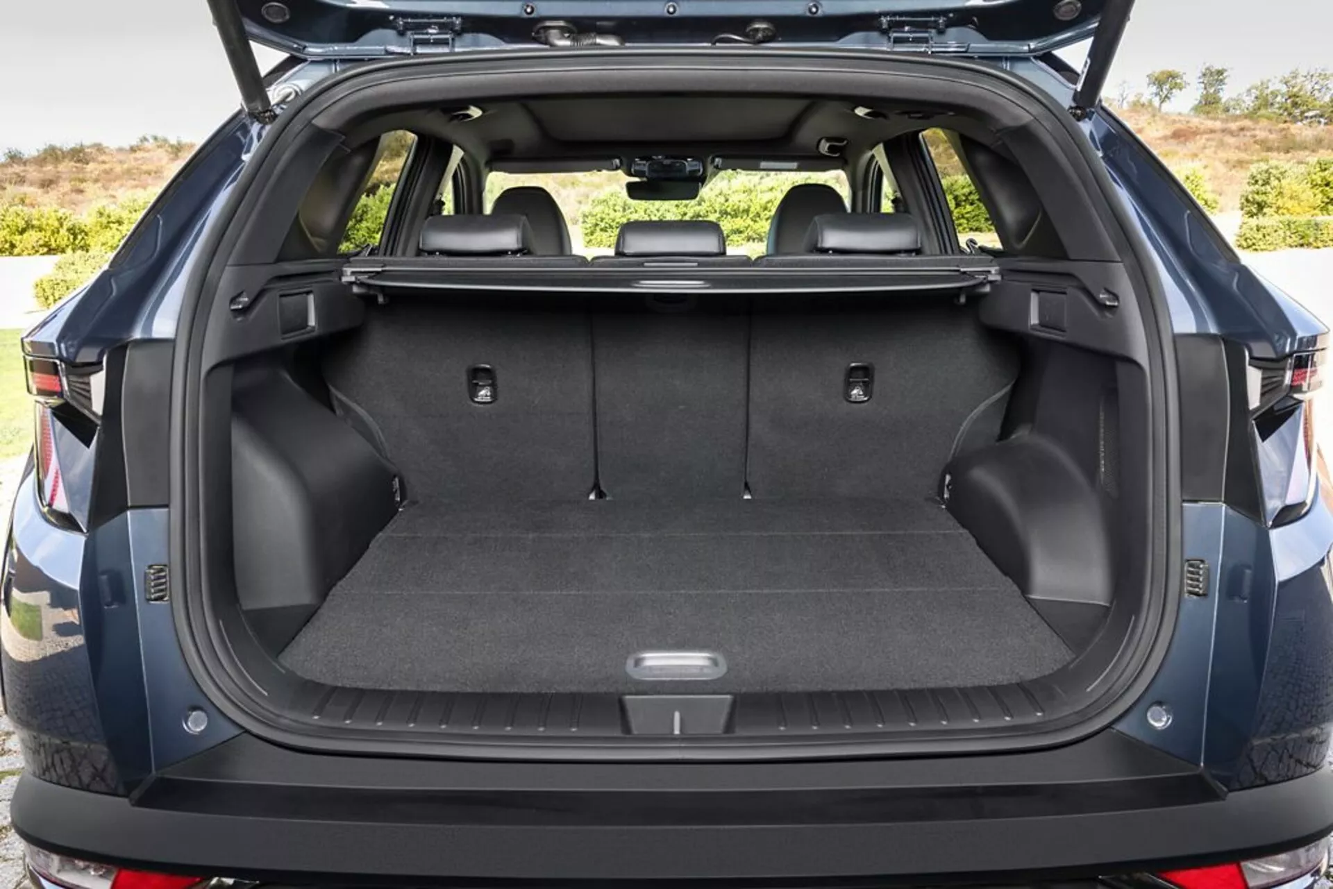 Zadný nákladový priestor Hyundai Tucson 2020 s otvoreným kufrom, ktorý predvádza podlahu nákladného priestoru pokrytú kobercom, body na upevnenie, úložné priestory, 12V zásuvku, sklopné zadné sedadlá, zasunutý kryt nákladu, odolné plastové bočné steny a náhľad na zadný nárazník, v exteriéri.