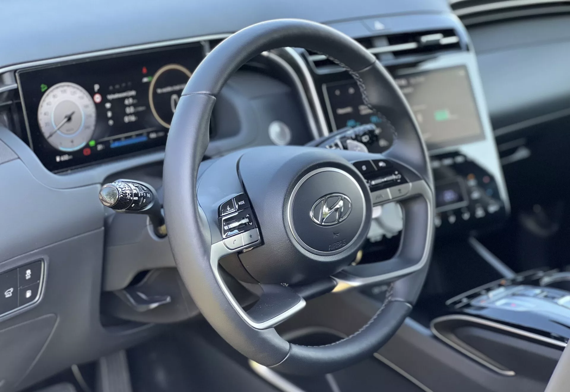 Vnútorný pohľad na Hyundai Tucson 2020 so zameraním na volant a funkcie palubnej dosky z pohľadu vodiča, zdôrazňujúc trojramenný dizajn s ovládacími tlačidlami, digitálny prístrojový panel a čiastočný výhľad na stredovú konzolu a ovládanie klímy.