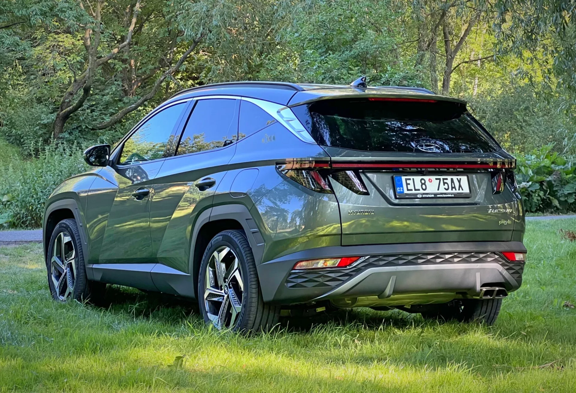 Zadný trojštvrtečný pohľad na Hyundai Tucson 2020 zdôrazňujúci LED zadné svetlá, parkovacie senzory, dvojitý výfuk, športový zadný spojler a strešné nosiče, zaparkovaný vonku na tráve predstavujúci aerodynamický dizajn a pokročilé funkcie moderného crossover SUV pre dobrodružstvo v prírode a praktické použitie.