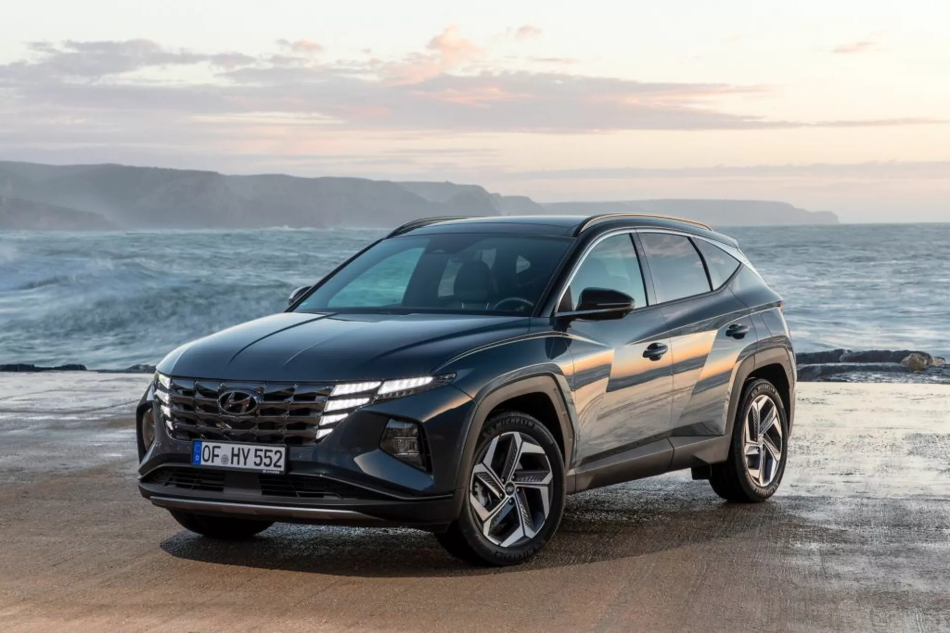 Moderné SUV Hyundai Tucson 2020 zaparkované na pláži s pozadím oceánu a horizontu, zvýrazňuje pohľad z prednej trojštvrte, ktorý zdôrazňuje dizajn vozidla, výraznú mriežku s logom, uhlové LED svetlá a športové zliatinové kolesá