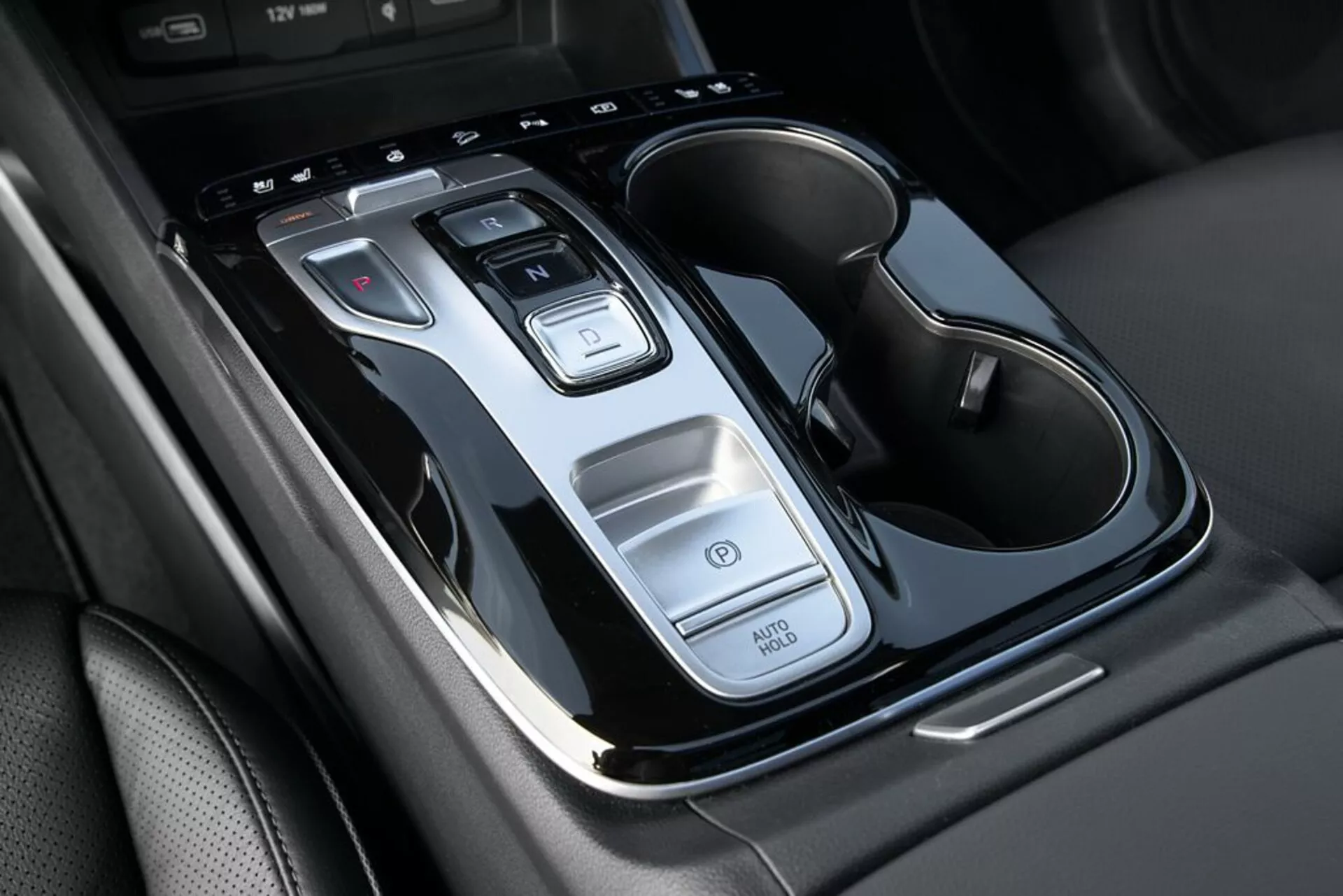 Detailný pohľad na stredovú konzolu Hyundai Tucson 2020 ukazujúci páku automatickej prevodovky, ovládacie tlačidlá, funkciu Auto Start/Stop, vypínač výstražných svetiel a dvojitý držiak na nápoje s chrómovými prvkami, z pohľadu zhora s miernym sklonom.