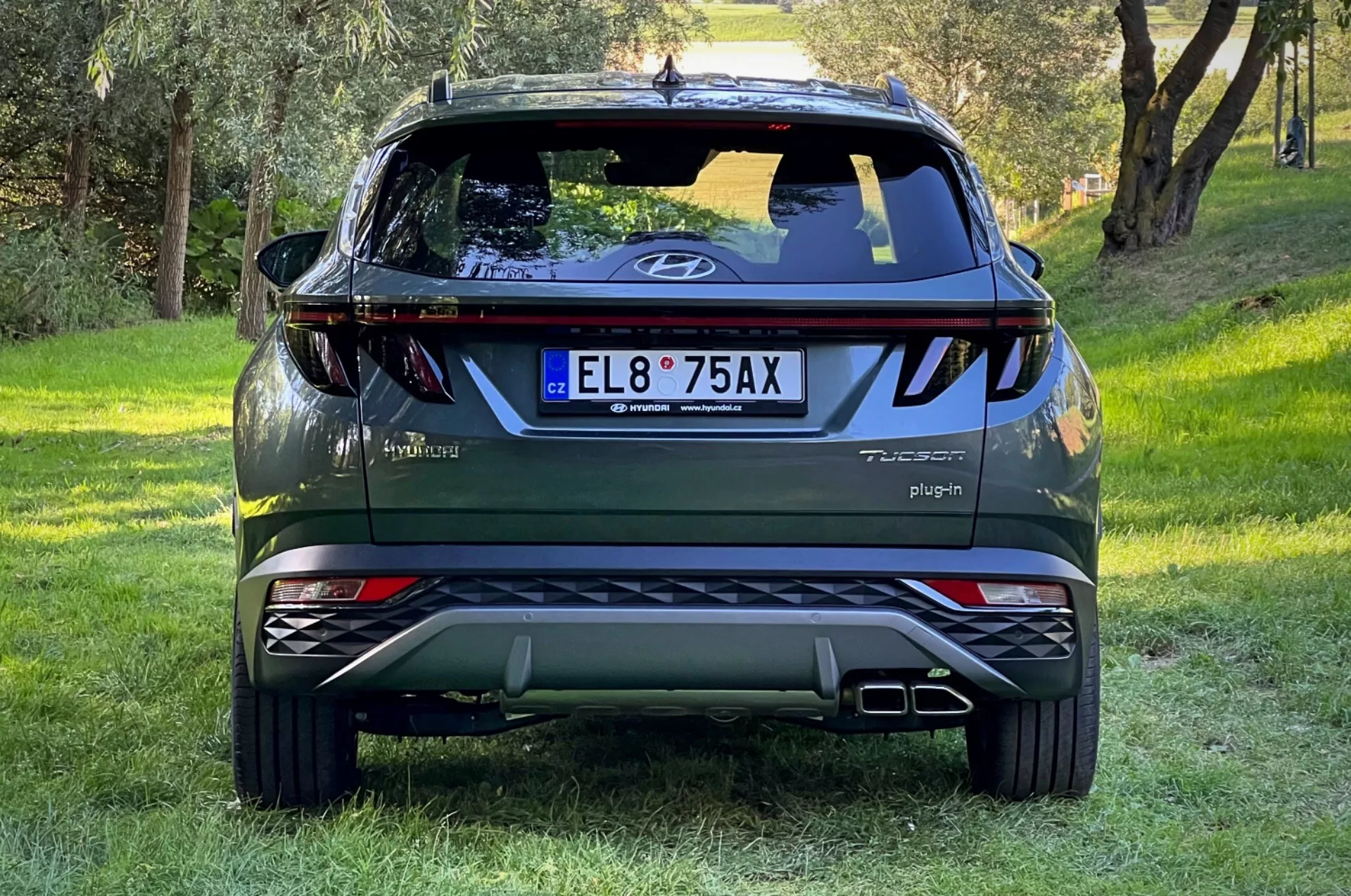 Zadný pohľad na SUV Hyundai Tucson 2020 na trávnom svahu s lesným pozadím, zobrazujúci logo na zadných dverách, odznaky Tucson a Hybrid, nápis Plug-in, odrazové svetlá, dynamické zadné svetlá, zadný stierač, vysoko umiestnené brzdové svetlo, dvojité výfukové koncovky, strieborné ochranné dosky a odznak AWD.