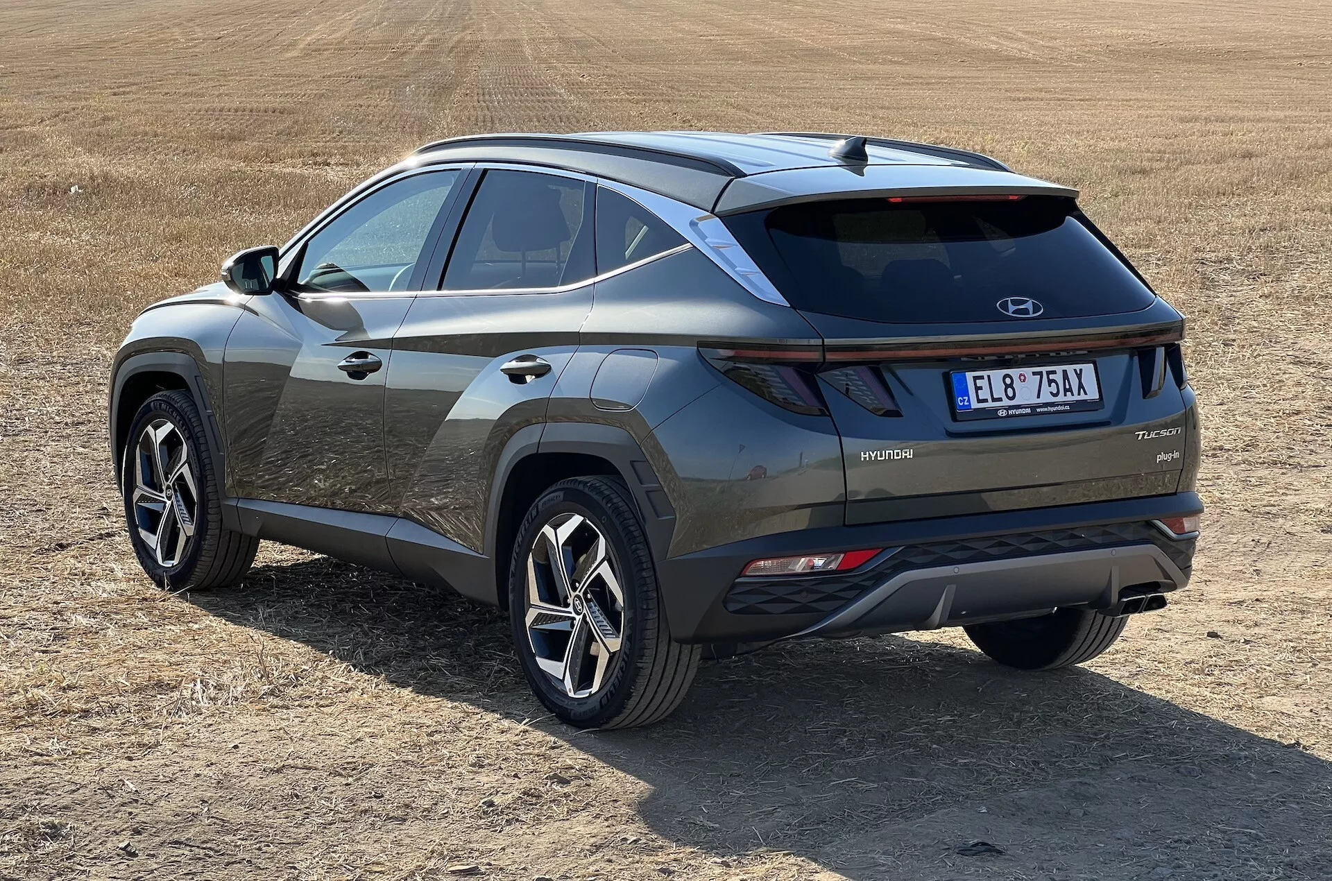 Moderné SUV Hyundai Tucson 2020 v šikmom pohľade ukazujúce zadné a bočné profily s označením hybridnej technológie na suchom travnatom poli, zdôrazňujúce LED zadné svetlá, kontrastnú vložku nárazníka a športové kolesá s viacerými lúčmi