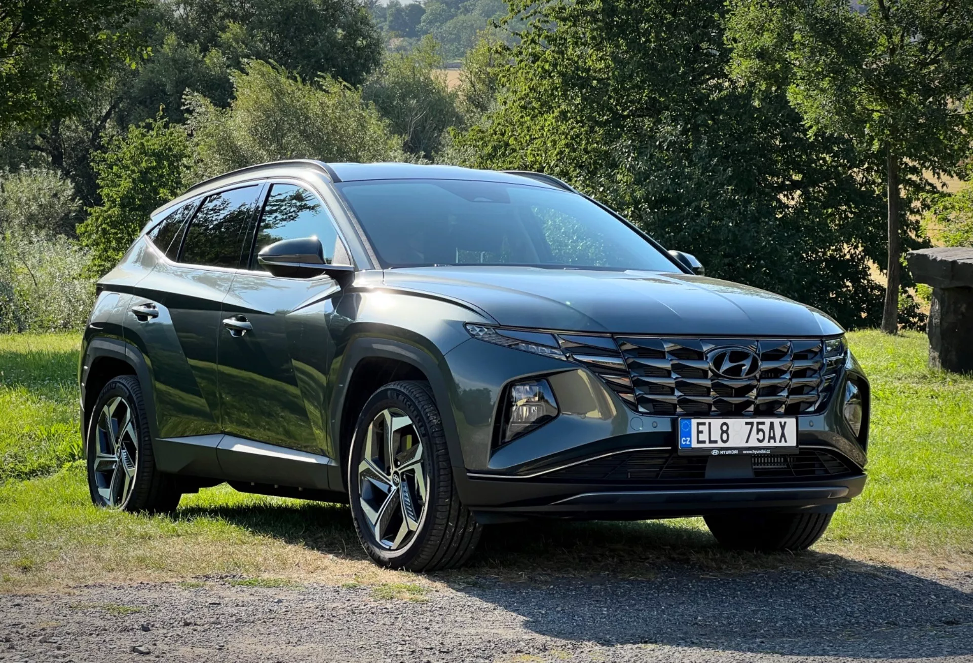 Hyundai Tucson 2020 zaparkované na tráve s vidieckym pozadím, zobrazujúce perspektívu troch štvrtín z prednej strany pre detailný pohľad na jej štíhle LED svetlomety, uhlovú prednú mriežku a lesklý tmavo farbený povrch