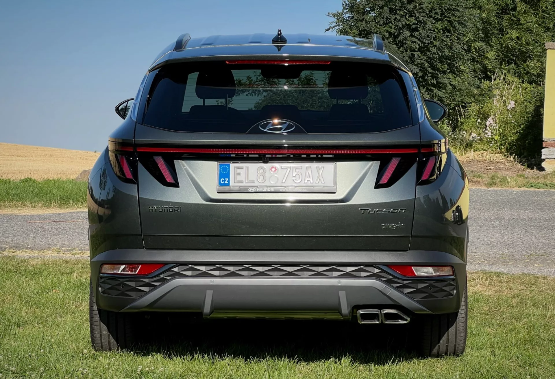 Zadný pohľad na Hyundai Tucson 2020 zobrazujúci horizontálne zadné svetlá, centrálny emblém, názov úrovne vybavenia, integrované parkovacie senzory na nárazníku a dvojitý výfukový systém, zaparkovaný na travnatom povrchu proti jasnému nebu
