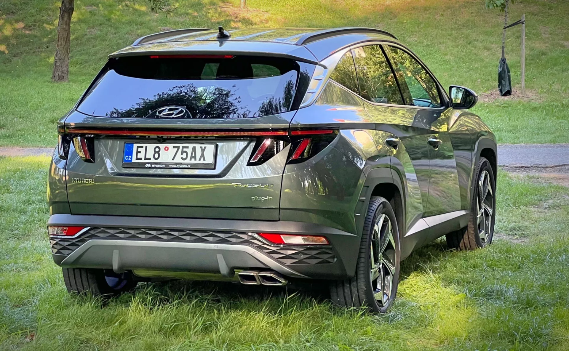 Zadný trojštvrtečný pohľad na Hyundai Tucson 2020, ktorý predvádza jeho LED zadné svetlá, aerodynamický dizajn so strešným spojlerom a anténou vo forme žraločej plutvy, prémiový tmavo zelený metalický finiš a vybavený veľkými zliatinovými kolesami a parkovacími senzormi, v prostredí crossover urban lifestyle