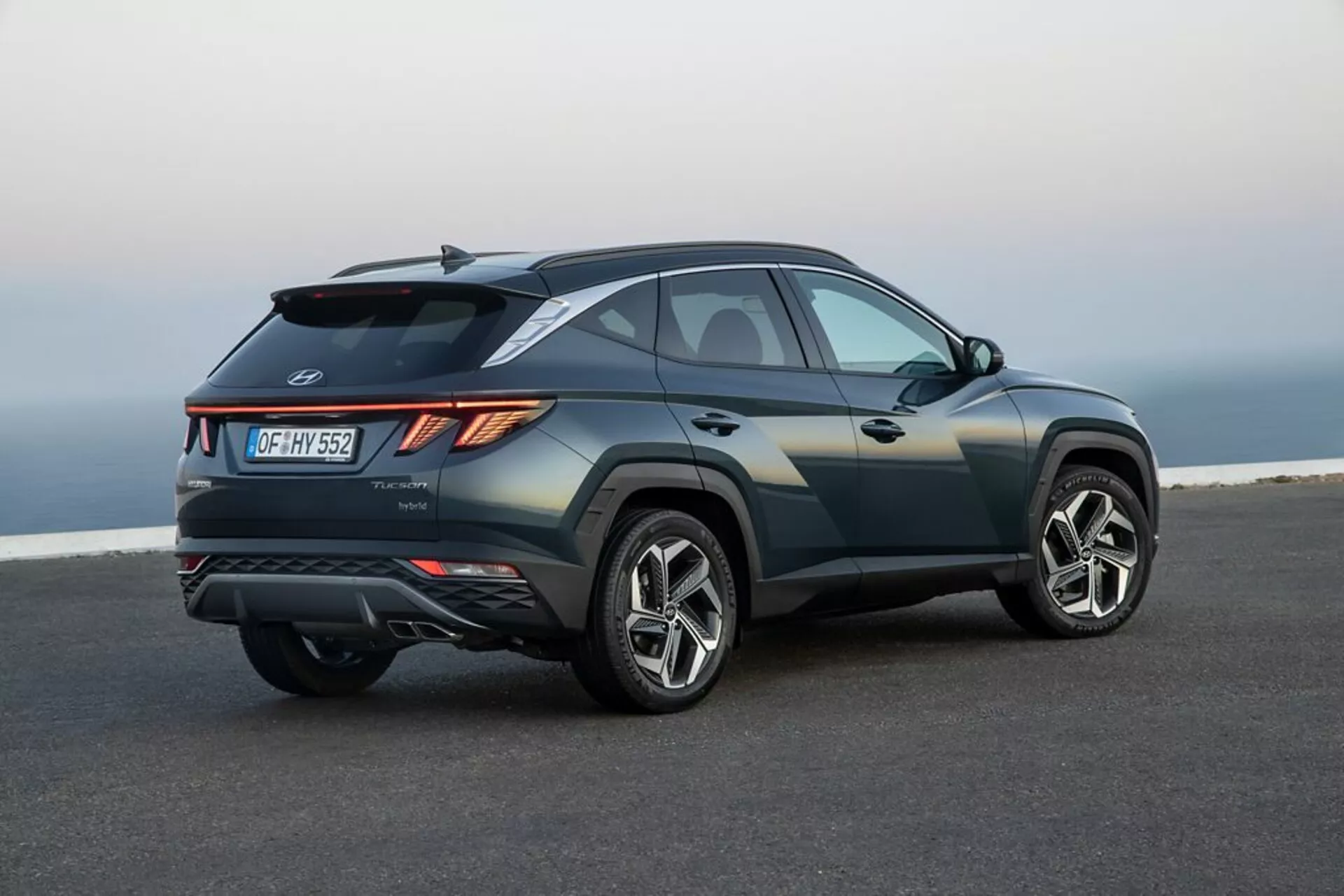 SUV Hyundai Tucson 2020 zaparkované na hladkom asfalte s pobrežnou alebo vyvýšenou krajinou na pozadí, ukazuje jeho elegantný, športový dizajn, rastúcu líniu opasku, sklonenú strechu, ostré ryhy na karosérii, uhlové LED zadné svetlá, zadný spojler, integrovaný difúzor, dvojité výfuky a veľké zliatinové kolesá