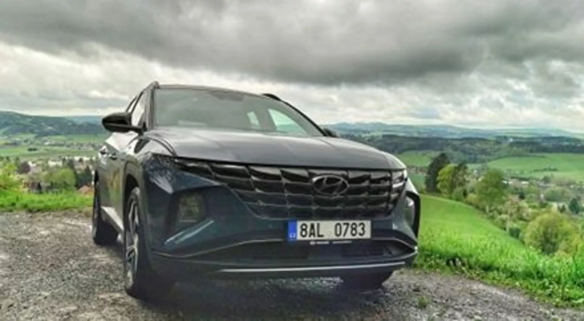 SUV Hyundai Tucson 2020 zaparkované na štrkovej ceste, ukazuje svoju odvážnu mrežu, štíhle LED svetlomety a metalický lak v dynamickom pohľade z nízkeho predného uhla, s bujnou kopcovitou krajinou a zamračeným nebom na pozadí