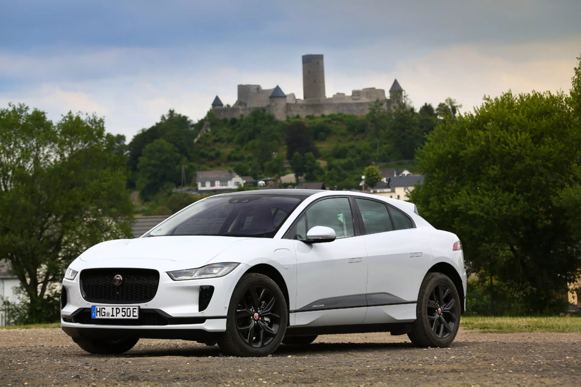 Biely crossover SUV Jaguar I-PACE so štyrmi dverami, rok 2018, zaparkovaný na štrku, ukazujúci svoj profil z trojštvrtečného predného uhla, vystavujúci jeho výraznú mriežku s emblémom, štíhle predné svetlomety zasahujúce do blatníkov, vysokovýkonné brzdové strmeňy viditeľné cez športové kolesá a zapustené kľučky dverí na pozadí historického hradu