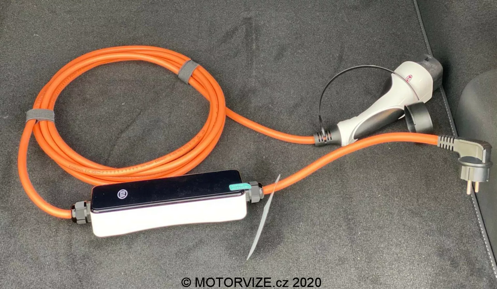 Nabíjací kábel a adaptér pre elektrické vozidlo (EV) Kia Ceed 2018, zahŕňajúci prenosný oranžový EVSE (Zariadenie na Zásobovanie Elektrickou Energiou Vozidiel) s vysokou viditeľnosťou pre bezpečnosť. Nastavenie obsahuje nabíjací konektor a zástrčku na použitie s domácou zásuvkou, ako aj riadiacu skrinku s LED indikátormi, všetko položené na koberci podlahy kufra alebo nákladného priestoru vozidla.