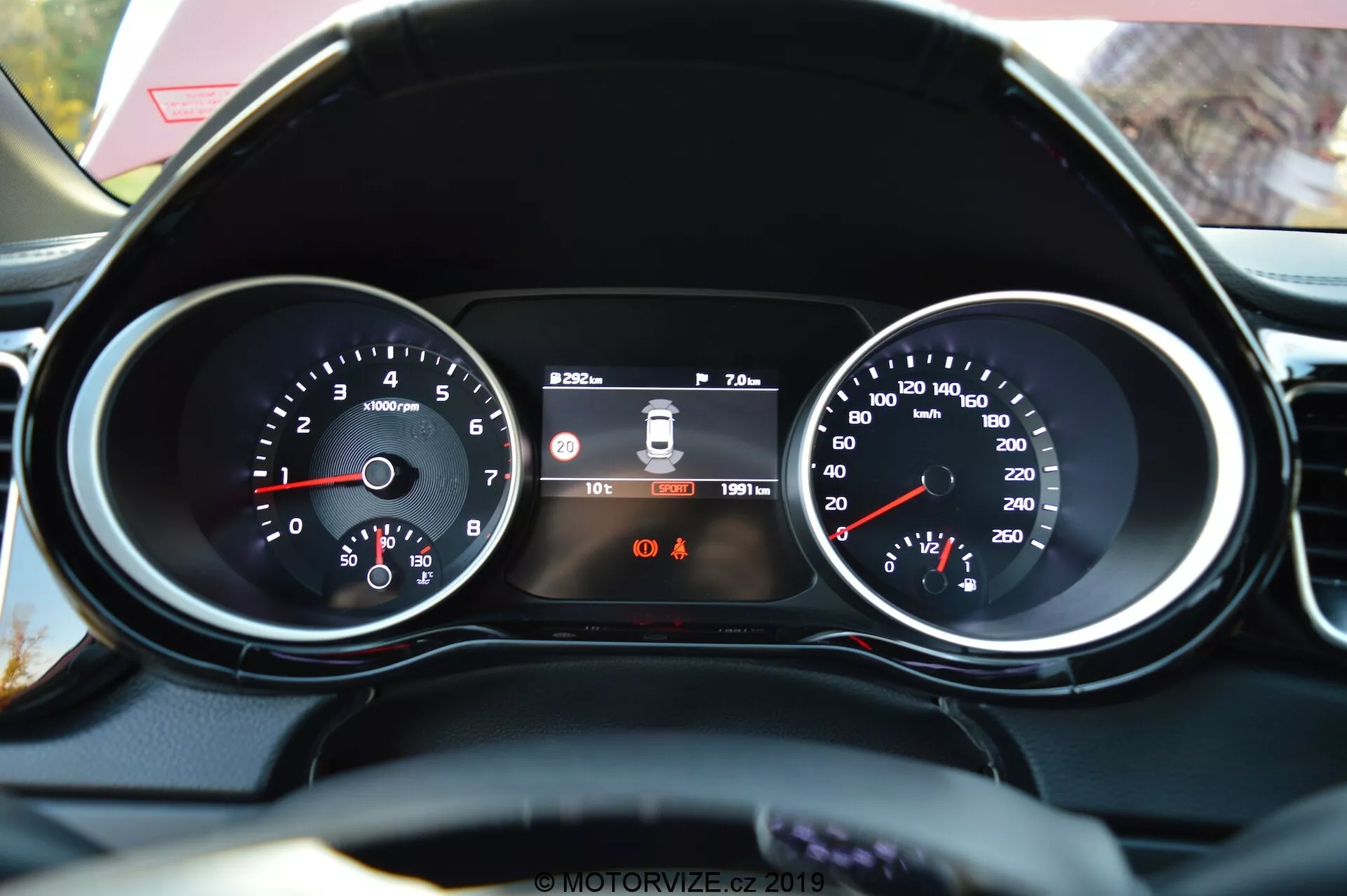 Detailný pohľad z pohľadu vodiča na prístrojový panel Kia Ceed 2021 a časť koženého volantu. Funkcie zahŕňajú ľavý otáčkomer s hodnotami RPM, centrálny digitálny displej zobrazujúci systém varovania pri opustení pruhu, čas a počítadlo kilometrov a pravý rýchlomer s rýchlosťou v km/h. Pod centrálnym displejom sú viditeľné dve výstražné kontrolky, pričom pripomienka zapnutia bezpečnostného pásu je identifikovateľná.