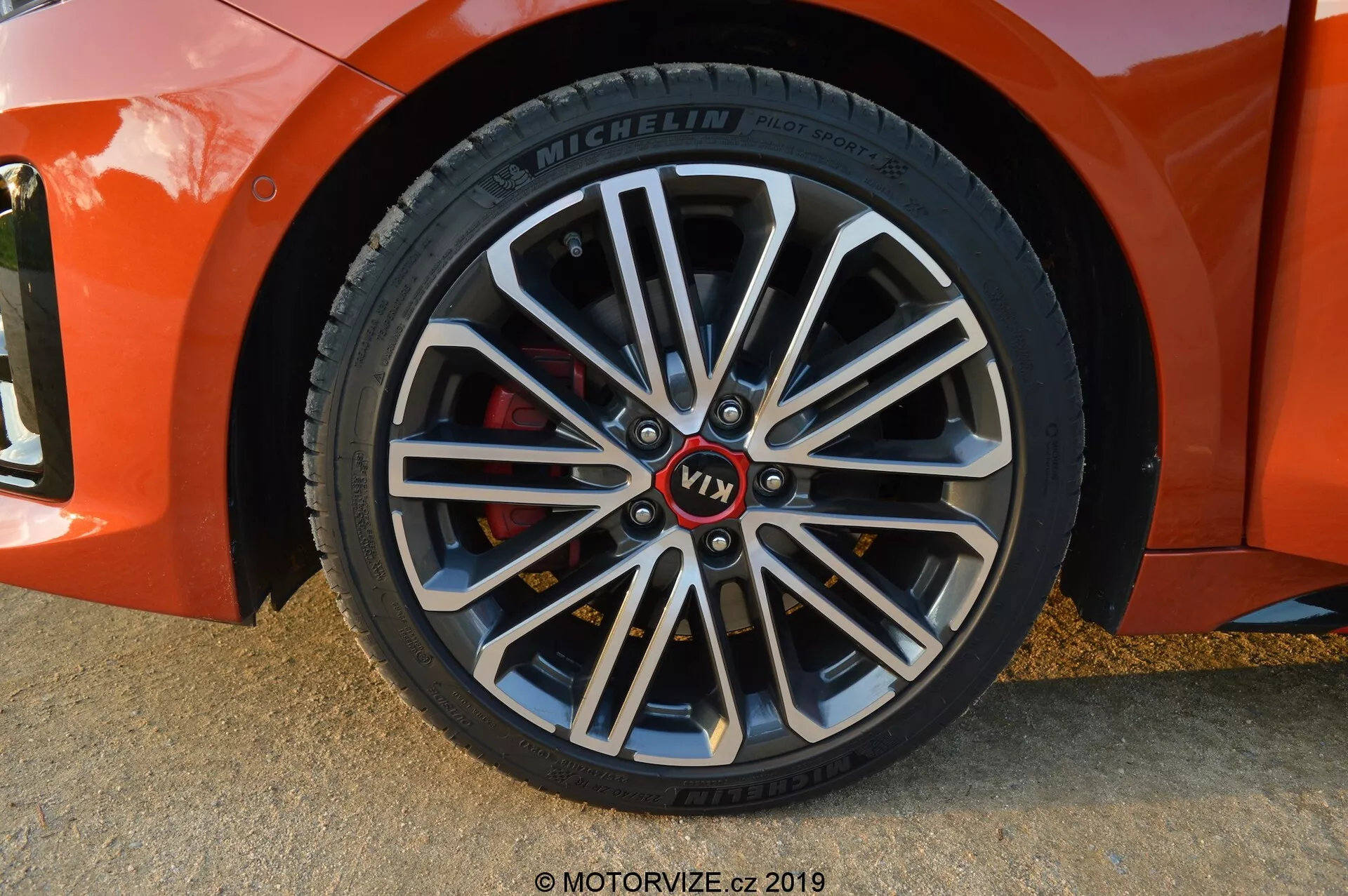 Detailný záber na predné koleso pasažiera Kia Ceed 2021, pod jemným diagonálnym uhlom, zobrazující viacramenné zliatinové koleso s tmavšími vnútornými časťami a jasnými opracovanými okrajmi. Obsahuje značkový náboj s červeným nápisom 'VT1', pneumatiku Michelin s podrobnými informáciami na bočnej stene a červený brzdový strmeň, ktorý signalizuje výkonovo orientované komponenty. Mäkké tiene a zvýraznenie naznačujú, že fotografia bola zhotovená v prirodzenom osvetlení, pravdepodobne počas zlatej hodiny.