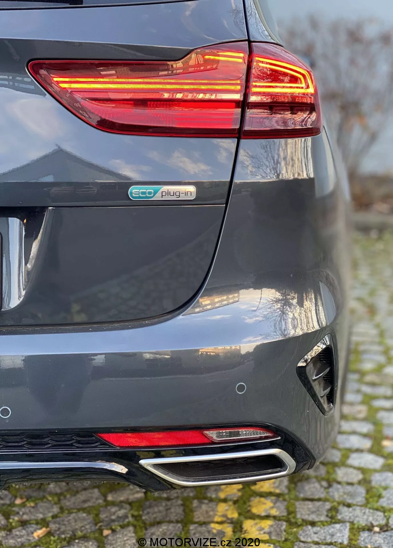 Detailný pohľad na zadnú časť Kia Ceed 2018 v metalickej šedej farbe, zvýrazňujúci osvetlené LED zadné svetlo, znak eco plug-in, tvarovaný zadný nárazník s reflektorom a difúzorom a dvojité chrómové koncovky výfuku. Perspektíva ukazuje spojenie zadného nárazníka a pravej strany, čím zdôrazňuje prvky hybridného elektromobilu a moderného dizajnu.