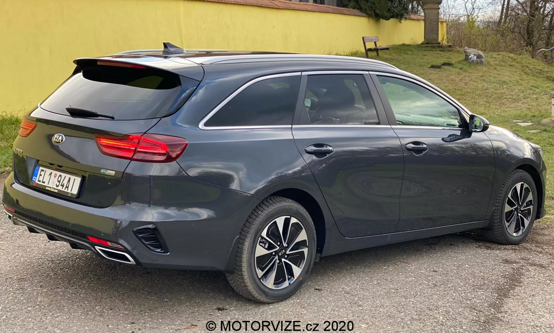 Tmavosivý metalický Kia Ceed 2018, zobrazený z trojštvrťového zadného pohľadu, zvýrazňujúci moderný hatchback alebo kombi dizajn so šikmým zadným oknom, integrovaným zadným spojlerom, viacramennými zliatinovými kolesami, kľúčkami dverí a spätnými zrkadlami v karosérii pravdepodobne s integrovanými smerovkami, charakteristický horizontálny zadný svetelný klaster pravdepodobne s LED technológiou, žraločia anténa, dvojité výfukové trubky, vysoko namontované tretie brzdové svetlo, odmrazovacia schopnosť zadného okna, senzory alebo odrazky naznačujúce parkovací asistenčný systém, fotografované zblízka s neutrálnym osvetlením.