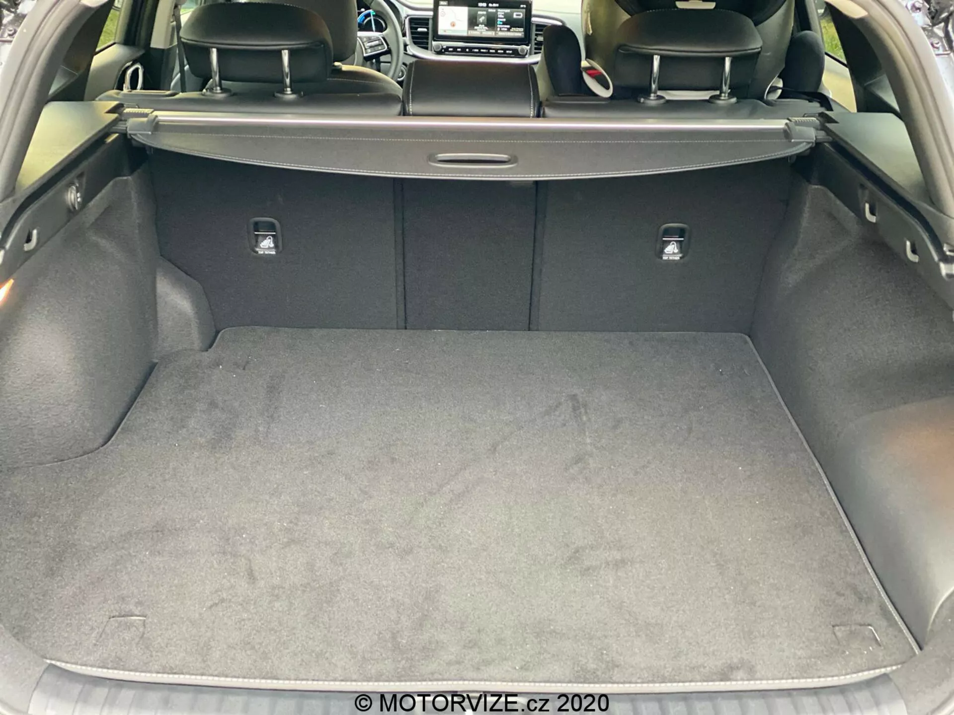 Detailný pohľad na batožinový priestor Kia Ceed 2018 zo zadnej perspektívy, zvýrazňujúc priestrannú a čistú nákladnú zónu s kobercovým povrchom. Dvere kufra sú zdvihnuté, odhaľujúc roletový kryt, operadlá zadných sedadiel tmavšej farby, pravdepodobne z kože, a strieborné úchyty na sklopenie sedadiel. Boky sú potiahnuté jemným materiálom a na pravej strane je viditeľná malá priehradka. Obrázok bol urobený z mierneho uhla, ukazujúc dizajn a materiály interiéru batožinového priestoru auta.