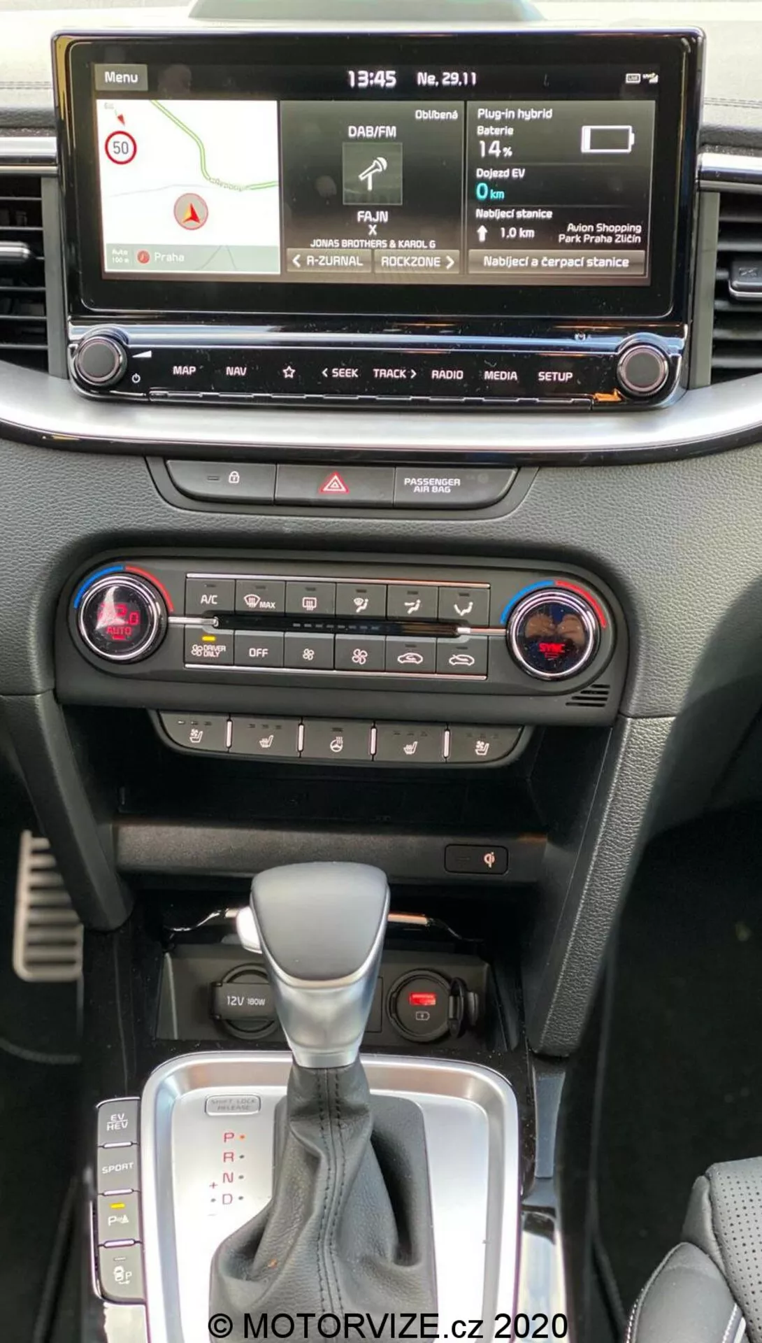 Interiér Kia Ceed 2018 zobrazujúci stredovú konzolu z pohľadu spolujazdca v prednom rade, s digitálnym infotainment displejom s navigačnou mapou, prehrávačom médií, nastaveniami vozidla, ovládaním klimatizácie s dvojzónovým systémom, tlačidlom výstražných svetiel, automatickou prevodovkou s koženým návlekom a slotom na kľúč bez kľúčového štartu.