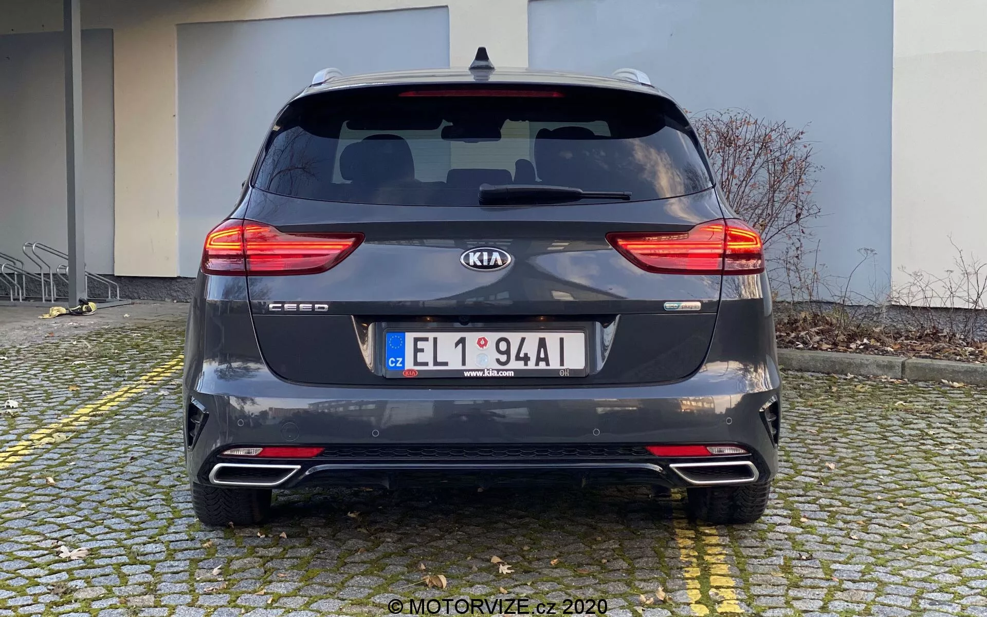 Zadný pohľad na Kia Ceed 2018 zaparkovanú na dláždenom povrchu blízko obrubníka. Auto, fotografované cez deň z priameho pohľadu na úrovni očí, ukazuje svoje rozdelené zadné LED svetlá, zadné okno s stieračom, emblém výrobcu, názov modelu, EČV, zadný nárazník s odrazovými prvkami a parkovacími senzormi, dvojitý výfukový systém a tmavosivú metalickú farbu. V pozadí je budova, čo naznačuje rezidenčnú alebo komerčnú zónu.