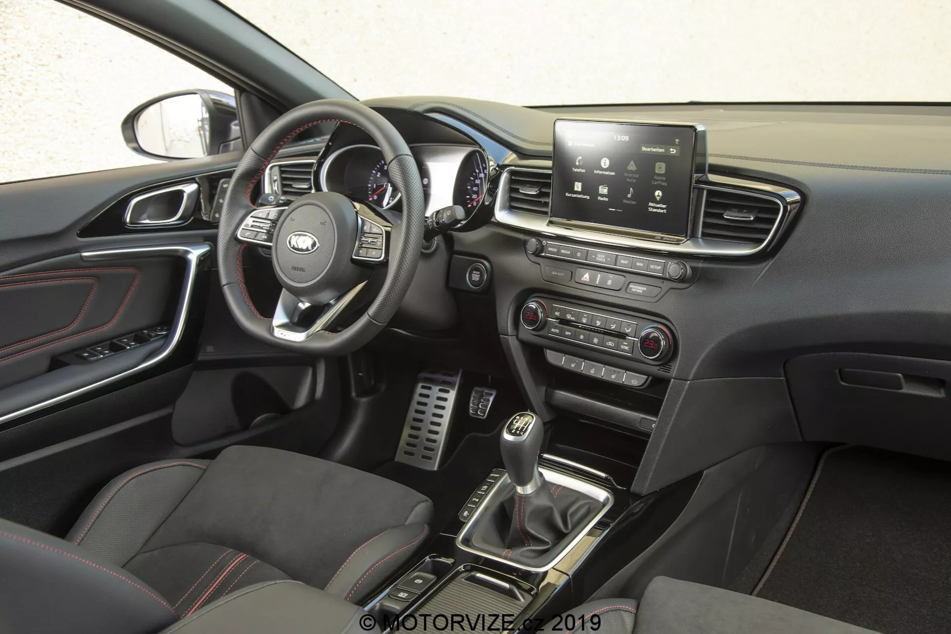 Interiér Kia Ceed 2021, zameraný na stranu vodiča z bočnej perspektívy. Volant s rozpoznateľným logom výrobcu obsahuje multifunkčné ovládače na správu infotainmentu, tempomatu, telefonického pripojenia a možno aj hlasových príkazov. Za volantom sú vidieť rýchlomer, otáčkomer a merače informácií pre vodiča. Stredová konzola má dotykovú obrazovku infotainmentu, ktorá ponúka navigáciu, nastavenie vozidla, prehrávanie médií a pripojenie Bluetooth. Nižšie sú manuálne ovládače pre audio a klimatizačné systémy. Radenie manuálnej prevodovky, obklopené tlačidlami pre jazdné režimy alebo kontrolu trakcie, a športové sedadlá s koženými a textilnými vložkami zvyšujú prémiový, výkonnostne orientovaný pocit. Kovové pedále v priestore pre nohy vodiča naznačujú dôraz na výkonnosť, zatiaľ čo detaily s chrómovým povrchom na ventilačných otvoroch, kľukách dverí a ďalších detailoch pridávajú sofistikovanosť. Dverový panel obsahuje ovládače na elektrické okná a možno aj ovládače elektrických zrkadiel, a je viditeľná časť spätného zrkadla, ktorá naznačuje funkcie ako automatické stmievanie alebo zabudovaný kompas.