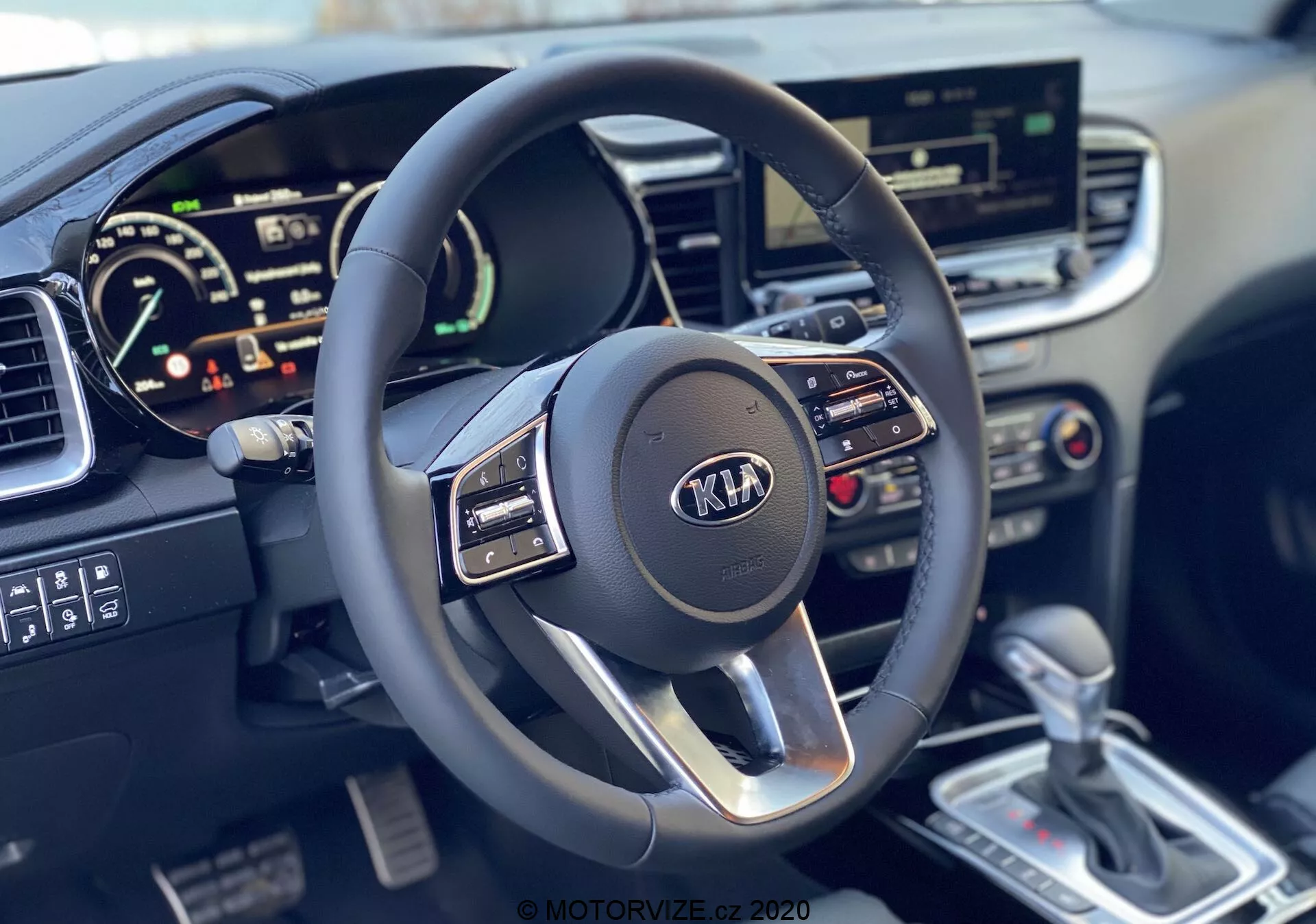 Pohľad z prvej osoby na interiér Kia Ceed 2018 s moderným trojlúčovým multifunkčným volantom s logom Kia a digitálnym prístrojovým štítom, zobrazujúcim tachometer a možný otáčkomer, dotykovým informačným systémom, ovládaním klimatizácie, striebornou automatickou radiacou pákou a detailným stredovým panelom.