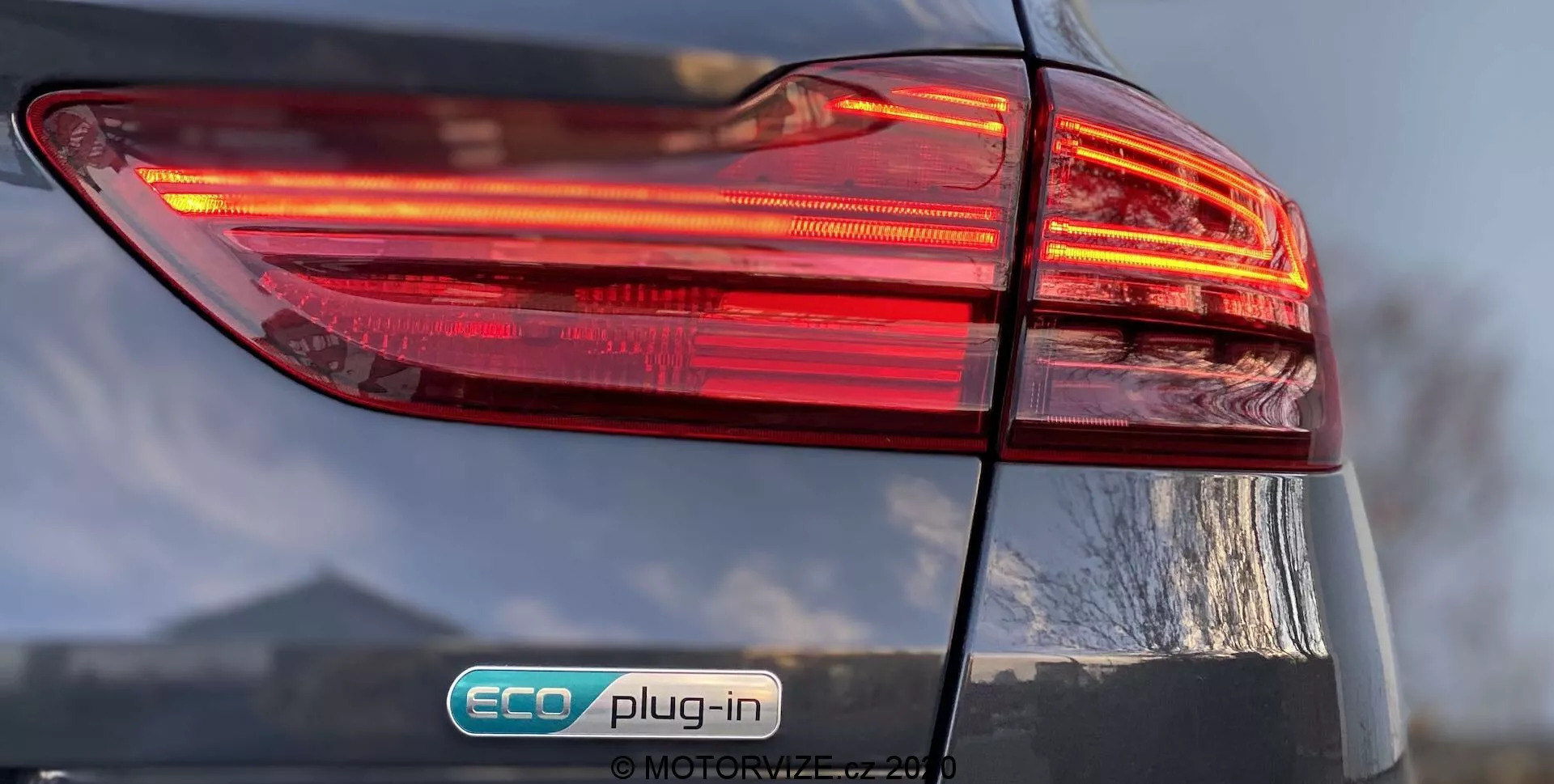 Detailný pohľad na zadnú ľavú stranu Kia Ceed 2018, zameraný na osvetlený zadný svetelný klaster s viacvrstvovým dizajnom. Odznak „ECO plug-in“ naznačuje, že ide o plug-in hybridné vozidlo. Lesklý povrch a čiastočne viditeľné veko kufra pridávajú detaily do celkového dizajnu vozidla.
