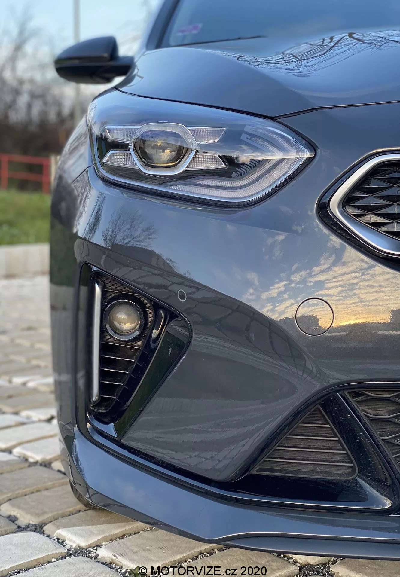 Detailný uhlový pohľad na prednú ľavú stranu tmavej Kia Ceed 2018, zvýrazňujúc elegantný dizajn predných svetiel s LED denným osvetlením, mriežku vo forme voštiny, zostavu hmlových svetiel a aerodynamický predný nárazník, s mäkkým rozmazaným pozadím a odleskom na lesklom laku.