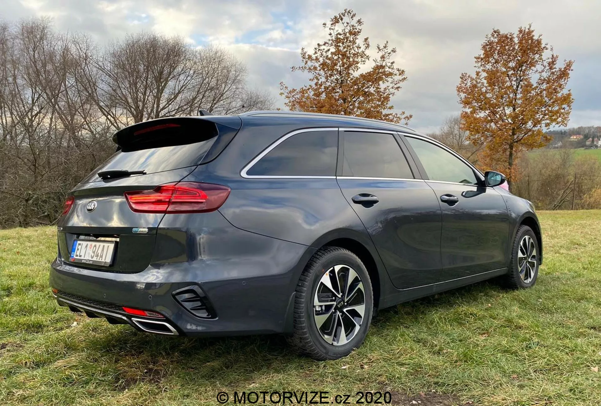Trojštvrťový zadný pohľad na moderný hatchback alebo kombi vozidlo, ktoré zobrazuje tmavosivý povrch, vertikálne štruktúrované zadné svetlá, dvojitý výfukový systém a viacramenné zliatinové kolesá, zaparkované na trávnatej ploche s miernym sklonom, obklopené stromami a zamračenou oblohou.