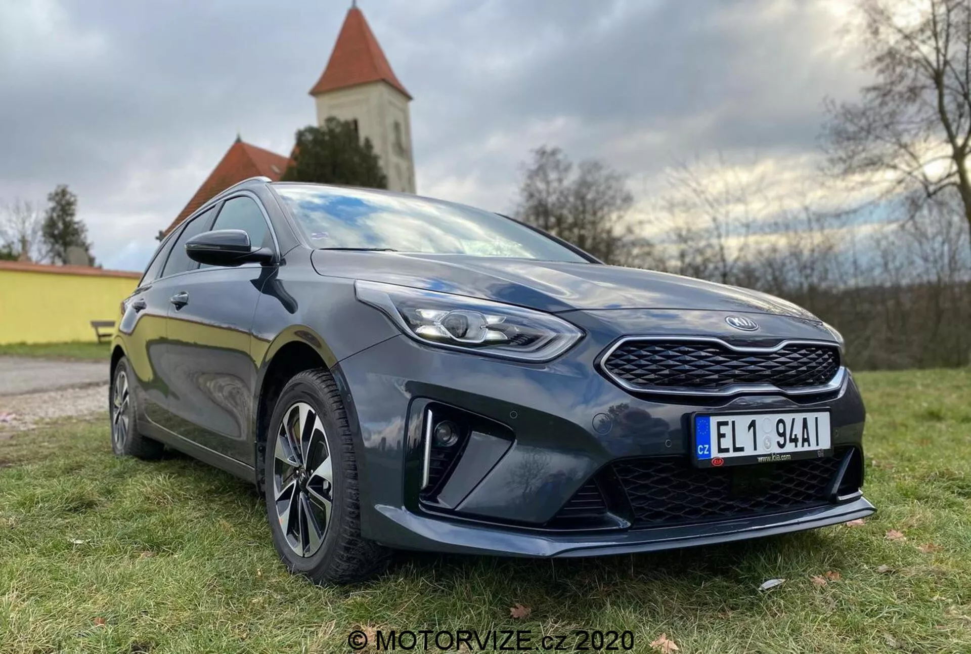 Detailný pohľad na moderný hatchback Kia Ceed 2018, fotografovaný z nízkeho predného trojštvrtového uhla, ukazujúci výraznú mriežku, ostré LED svetlomety a bočný profil až po dvere vodiča. Auto je tmavé, pravdepodobne tmavomodré alebo čierne, s lesklým leskom, ktorý vyzdvihuje jeho dobrý udržiavaný stav. Významné dizajnové prvky zahŕňajú vzorovanú prednú mriežku, ostré svetlomety s čírymi šošovkami, výrazné aerodynamické prvky na dolnom prednom nárazníku, viaclúčové zliatinové kolesá, bočné zrkadlá vo farbe karosérie s integrovanými smerovkami a športový bočný prah. Stance auta naznačuje rovnováhu medzi komfortom a športovosťou. Prirodzené denné svetlo s mäkkým osvetlením zvyšuje detaily a v pozadí je viditeľná prírodná krajina s kostolom alebo vežou.