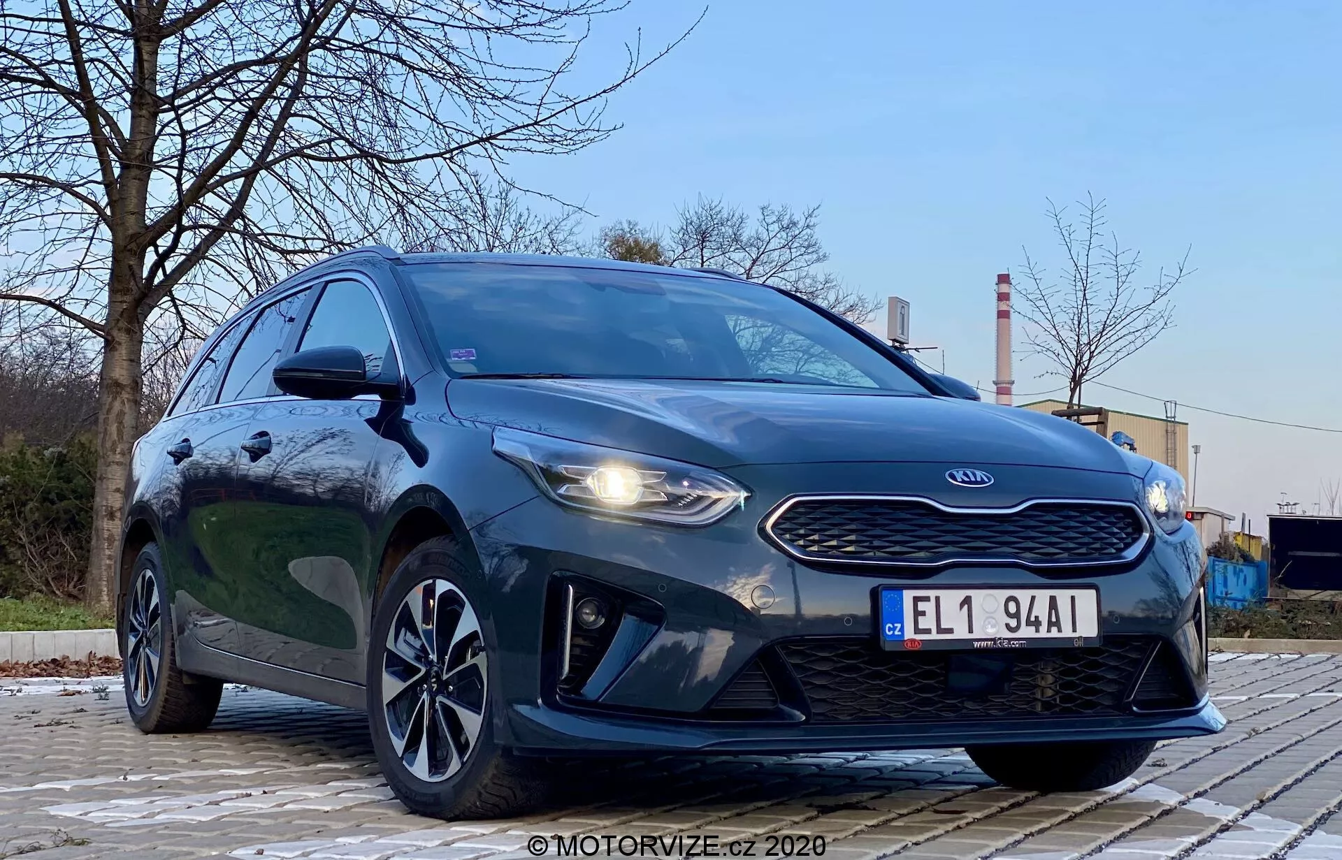 Fotografia Kia Ceed 2018, urobená z nízkeho, predného ľavého uhla, zvýrazňujúc masku, svetlomety a dizajn prednej časti vozidla. Agresívny štýl auta je zvýraznený výraznými svetlometmi, maskou v tvare tigrieho nosu a kontúrami na kapote. Záber ukazuje aj LED denné svetlá, aerodynamický predný nárazník a štýlové zliatinové kolesá. Prostredie je vonkajšie, so stromami a priemyselnými štruktúrami v pozadí, čo poskytuje kontrast. Tmavá, metalická farba vozidla je lesklá a dobre osvetlená, s odrazmi naznačujúcimi čistý povrch. Obraz sa zameriava na auto s dramatickou perspektívou z nízkeho uhla, za mäkkého vonkajšieho osvetlenia.