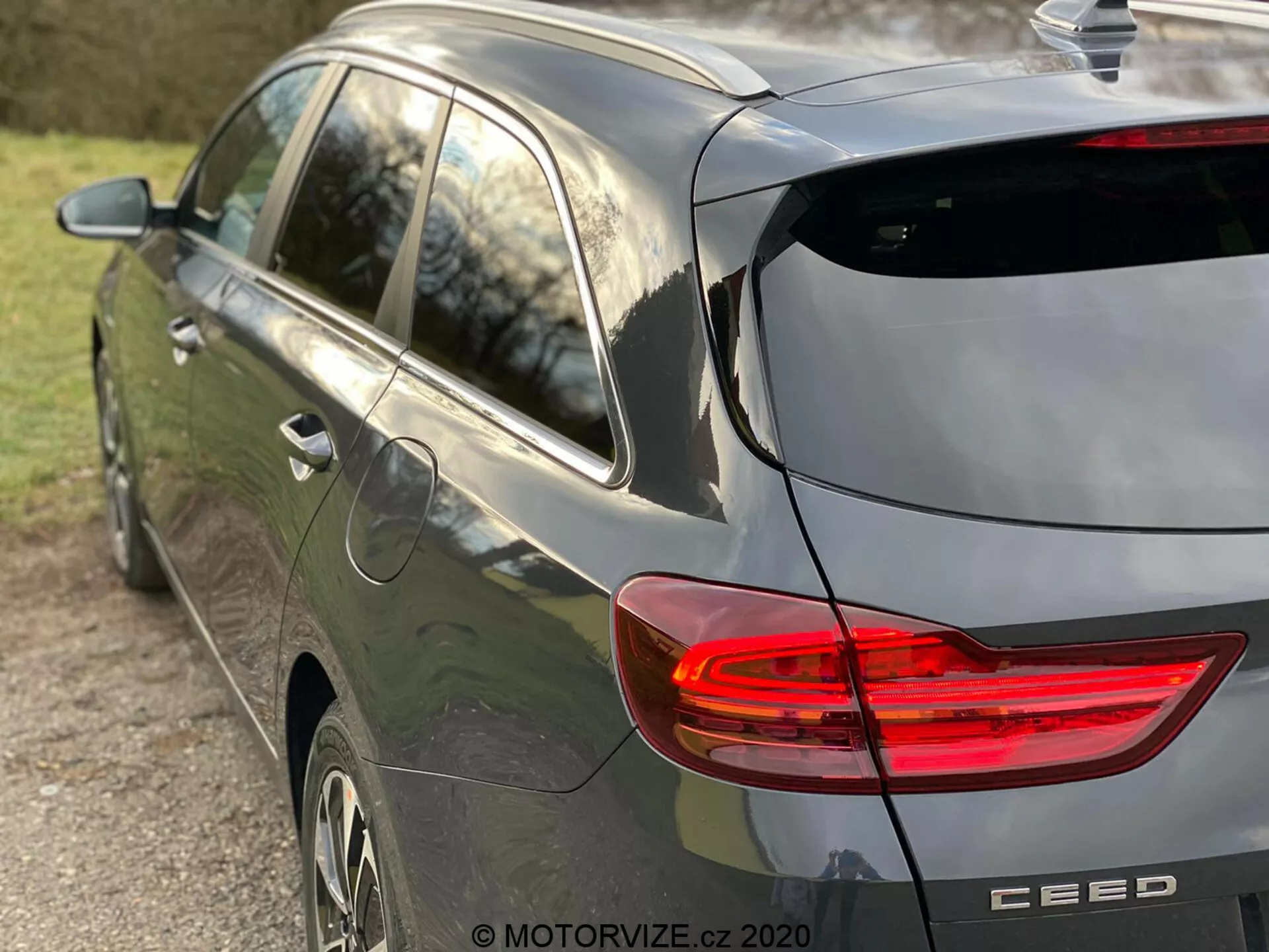 Čiastočný pohľad z boku a zozadu na moderný šedý hatchback Kia Ceed 2018, zdôrazňujúci osvetlené zadné svetlo v tvare písmena C, dizajn hatchbacku so zadným stieračom a jemným strešným spojlerom, anténu v tvare žraločej plutvy, hliníkové kolesá a značkový odznak s názvom modelu 'CEED', zachytené v prirodzenom neskorom popoludňajšom svetle a s malou hĺbkou ostrosti so zameraním na vozidlo.