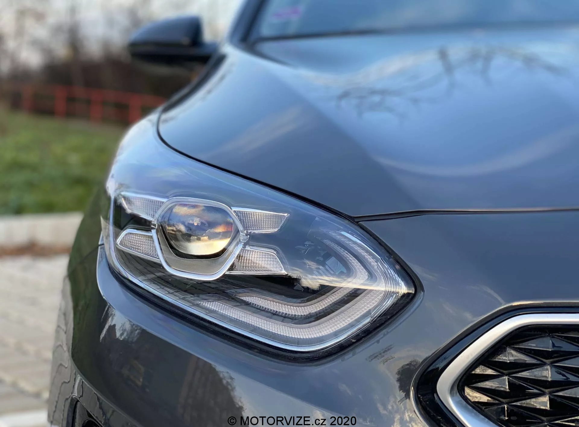 Detailný záber na prednú ľavú časť Kia Ceed 2018, zameraný na sofistikovaný svetlomet a časť prednej mriežky. Záber je robený z nízkeho uhla, čo zvýrazňuje elegantný a moderný dizajn vozidla s viditeľnou pokročilou osvetľovacou technológiou, pravdepodobne vrátane LED a adaptívnych svetlometov. Rozmazané pozadie zvýrazňuje aerodynamické prvky a luxusný vzhľad vozidla, čo naznačuje výkonovo orientované vozidlo.