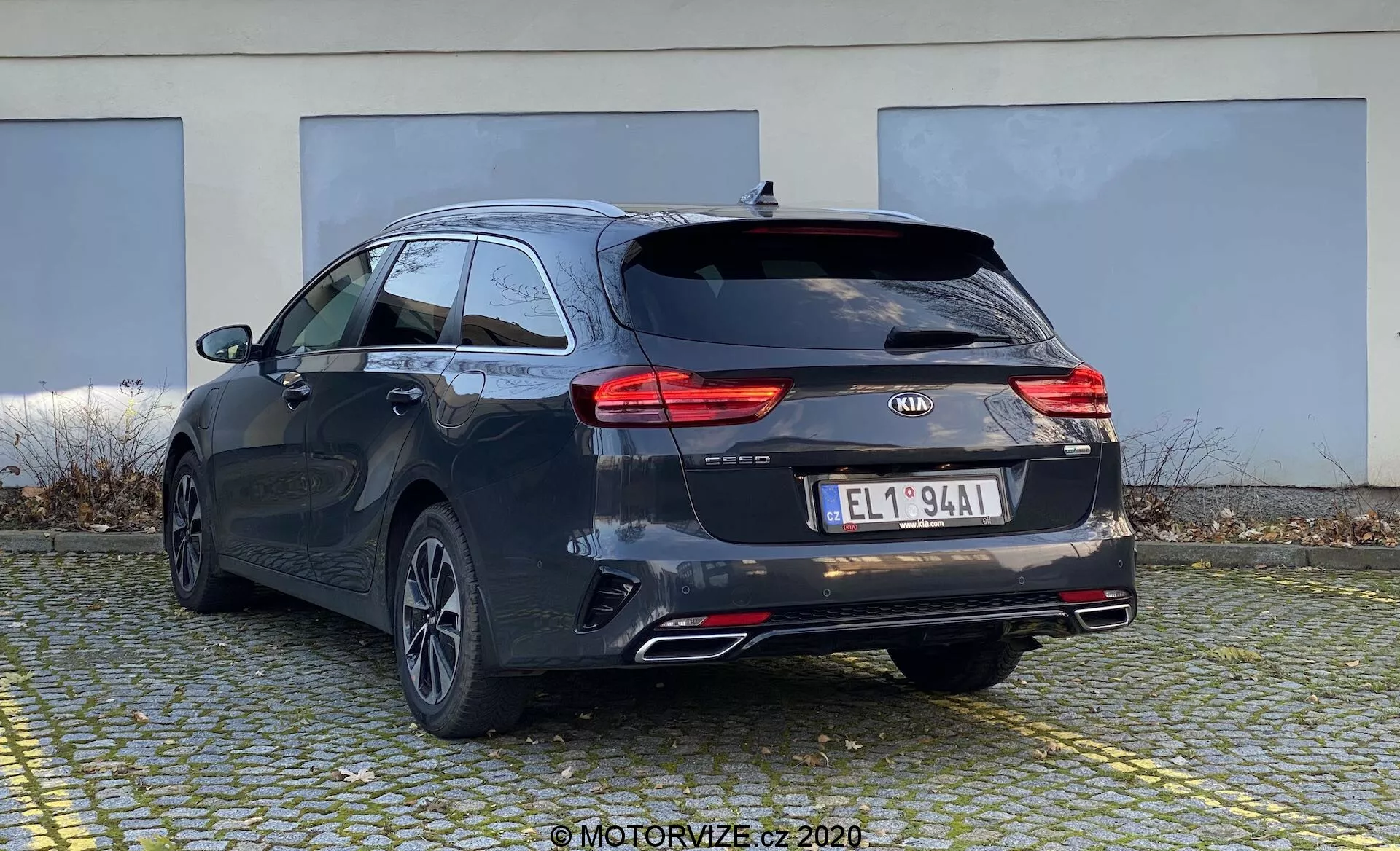Obrázok zobrazuje Kia Ceed 2018 z trojštvrťového zadného pohľadu, vrátane zadnej a bočnej časti kombi. Vozidlo má tmavý odtieň, pravdepodobne čierny alebo tmavosivý, s charakteristickými líniami a mierne sa zvažujúcou strechou pre športový vzhľad. Viditeľné sú moderné LED zadné svetlá, logo Kia, názov modelu 'CEED', ďalšie označenia, európska registračná značka, zadné kolesá s viaclúčovými zliatinovými ráfikmi, dvojité výfukové potrubie a zadný stierač. Pravdepodobne je vybavený zadnou kamerou a parkovacími senzormi, ktoré nie sú viditeľné. Malý strešný spojler a strešné lišty naznačujú možnosti pre prídavný náklad. Auto je zaparkované na dláždenej ploche s budovou v pozadí, čo zachytáva jeho zadný a časť bočného profilu z mierne nízkeho uhla.