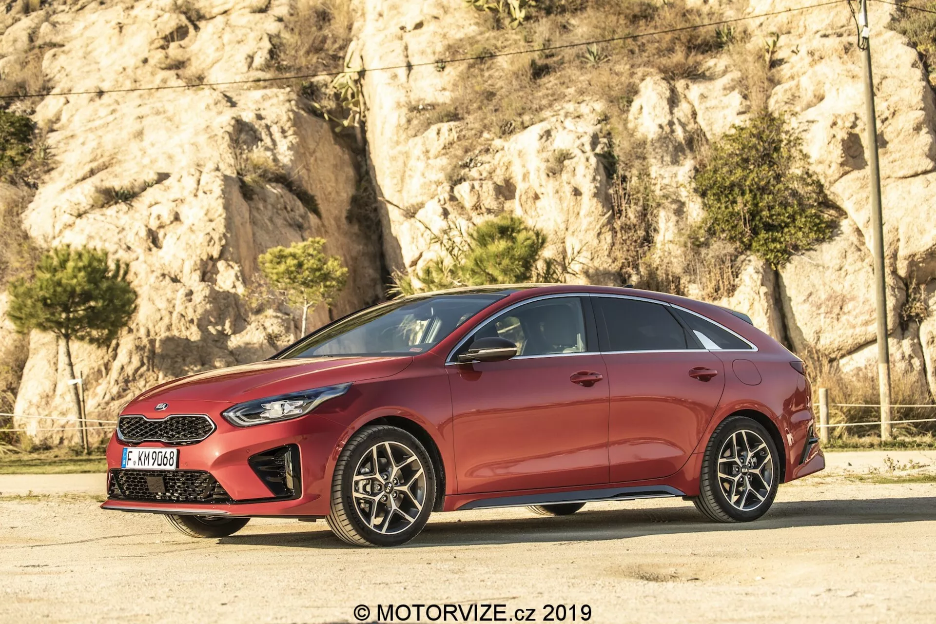 Bočný profil červeného hatchbacku Kia Ceed 2021, s elegantným, aerodynamickým dizajnom so sklónenými líniami strechy a kontúrovanými líniami na dverách. Vozidlo, päťdverový model, má chrómové okenné lemovanie a začiernené B-stĺpiky, vytvárajúce efekt nepretržitého skleneného panelu. Viaclenové zliatinové kolesá s viditeľnými kotúčovými brzdami dodávajú športový nádych. Ostré svetlomety a čiastočne viditeľná mriežka značia predok, zatiaľ čo zrkadlá vo farbe karosérie s integrovanými smerovými svetlami zlepšujú viditeľnosť a bezpečnosť.