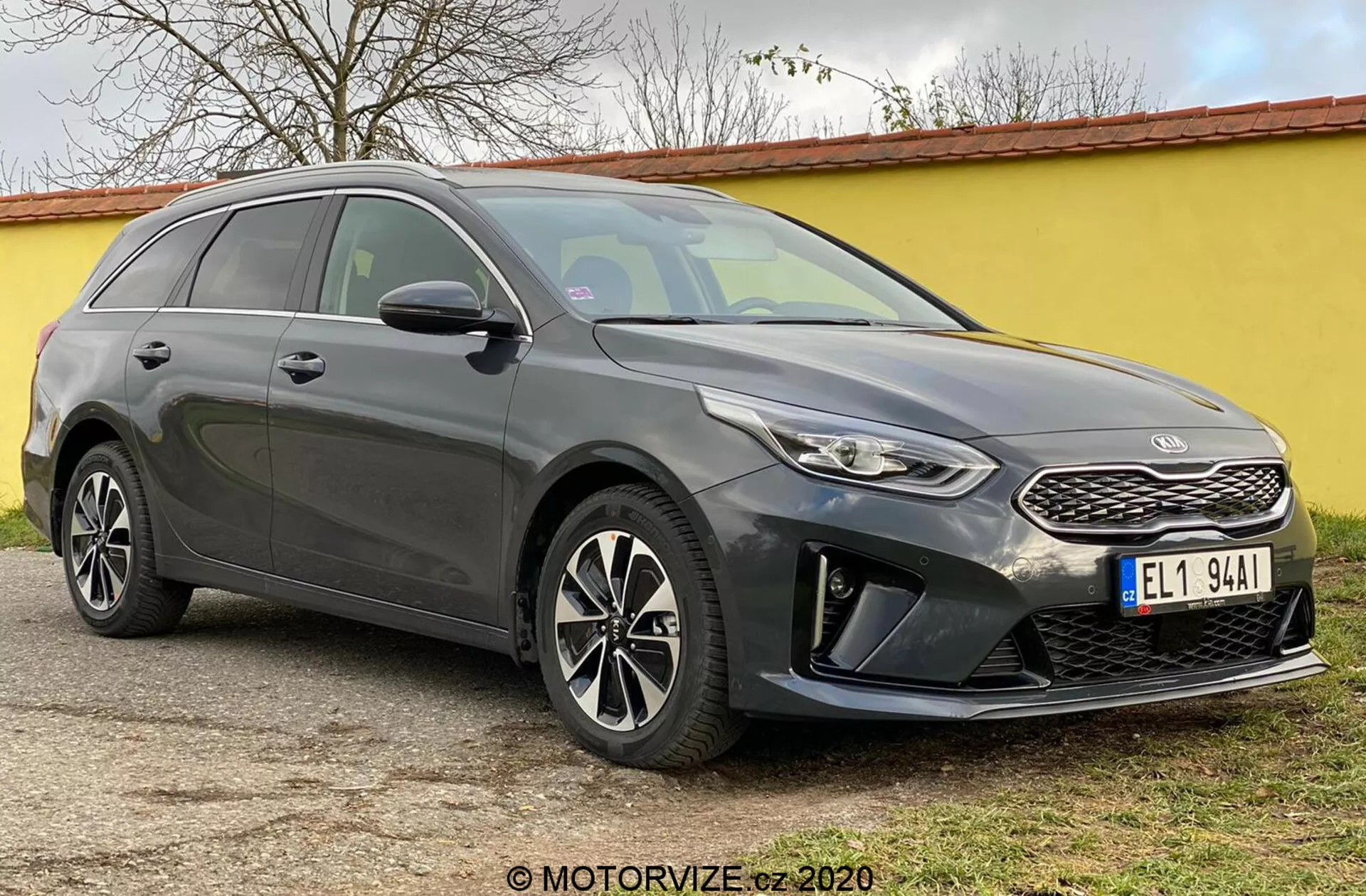 Obrázok ukazuje Kia Ceed 2018, moderné kombi zachytené z uhla prednej tri štvrtiny, zdôrazňujúce jeho prednú časť, vrátane štruktúrovanej masky chladiča s výrazným emblémom značky, úzke spätné svetlomety s LED denným osvetlením a predného nárazníka s krytmi hmlových svetiel a parkovacími senzormi. Elegantné línie kapoty vedú k prudko sklonenému čelnému sklu, čo zvýrazňuje aerodynamický profil auta. Viditeľné sú viaclúčové zliatinové kolesá a integrované smerové svetlá vo vonkajších zrkadlách, spolu s naklonenou strechou smerom dozadu a strešnými nosičmi, ktoré zdôrazňujú štýl aj úžitok. Tónované okná a línie karosérie po stranách pridávajú sochársky vzhľad. Auto je zastavené na rovine vonkajšej ploche.