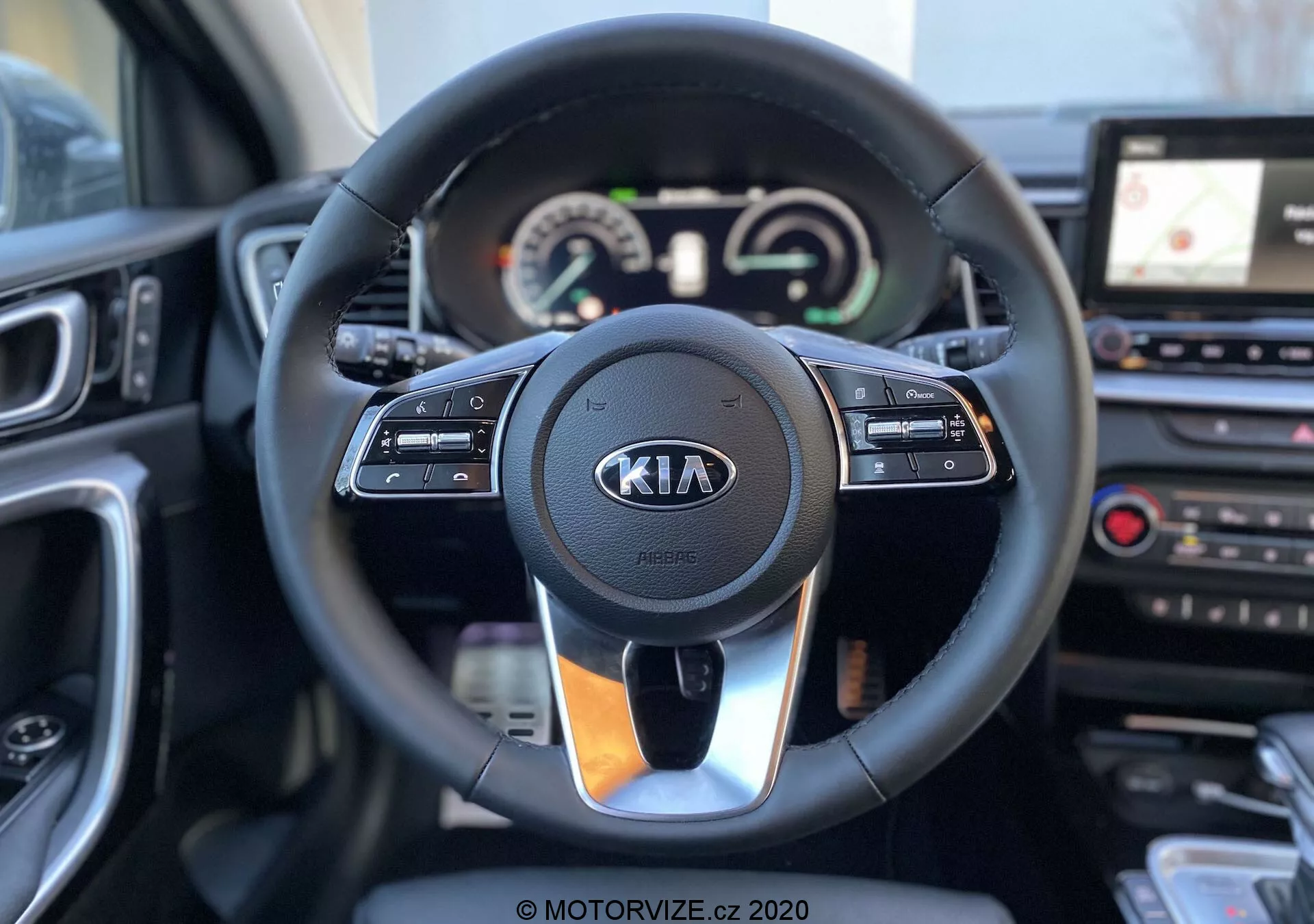 Interiér modelu Kia Ceed 2018 zameraný na volant a palubnú dosku z pohľadu sedacej polohy vodiča. Moderný volant obsahuje tlačidlá pre ovládanie tempomatu, multimédií a hands-free komunikácie, s výrazným logom Kia. Za volantom je prístrojový panel s analógovými ukazovateľmi a digitálnym displejom ukazujúcim rýchlomer a otáčkomer, vedľa multifunkčého displeja. Na pravej strane je viditeľná časť stredovej konzoly s dotykovým infotainment systémom a GPS navigáciou, ako aj tlačidlá ovládania klimatizácie. Obraz je ostrý, so zvýraznením volantu a jeho najbližšieho okolia.