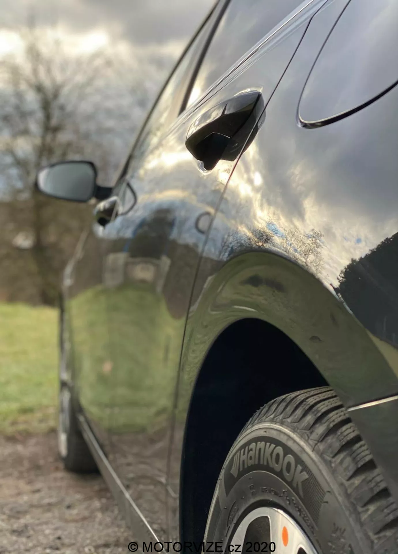 Detailný bočný pohľad na Kia Ceed 2018, zvýrazňujúci prvky ako zadné koleso s pneumatikou 'Hankook', aerodynamickú kľučku dverí a čiastočne rozmazané spätné zrkadlo. Fotografia je urobená z nízkeho uhla s malou hĺbkou ostrosti, ktorá ostro zobrazuje zadnú pneumatiku a spodnú časť dverí, zatiaľ čo pozadie je umelecky rozmazané. Auto má tmavú, lesklú povrchovú úpravu odrážajúcu oblohu a okolie, čo zdôrazňuje elegantné dizajnové línie a dobre udržiavanú karosériu.