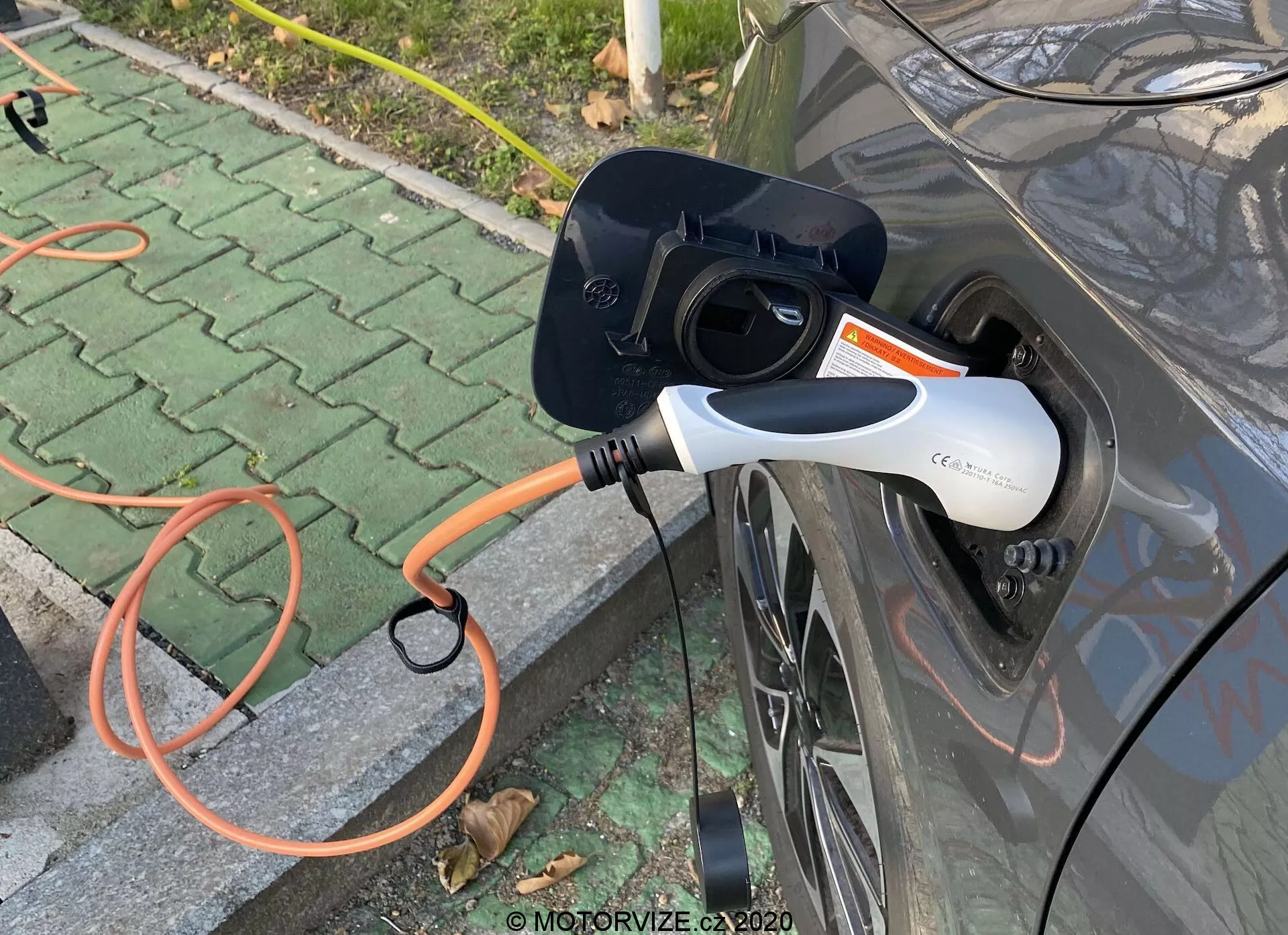Detailný bočný pohľad na sivé elektrické vozidlo (EV) počas nabíjania, zvýrazňujúci otvorený nabíjací port a pripojený oranžový EVSE konektor. Moderný biely a čierny nabíjací konektor má LED indikátor. Obrázok sa sústreďuje na proces nabíjania, zasadený do vonkajšieho prostredia s dláždeným povrchom a rozhádzanými listami.