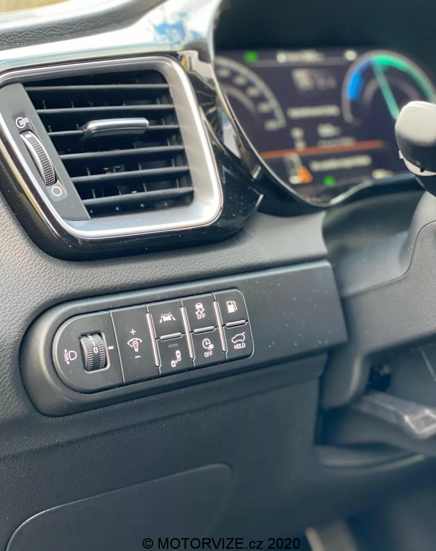 Detailný záber interiéru Kia Ceed 2018, ktorý ukazuje ovládacie tlačidlá pre nastavenie bočných zrkadiel, vyhrievanie zadného okna, vyhrievanie volantu, varovanie pred opustením jazdného pruhu, parkovacieho asistenta a trakčného systému. Obrázok tiež zobrazuje elegantný výduch klimatizácie, časť digitálneho prístrojového panela v rozmazanom pozadí a okraj volantu. Interiérové materiály sú prevažne z tvrdých plastov s matnou povrchovou úpravou, zachytené na dennom svetle.