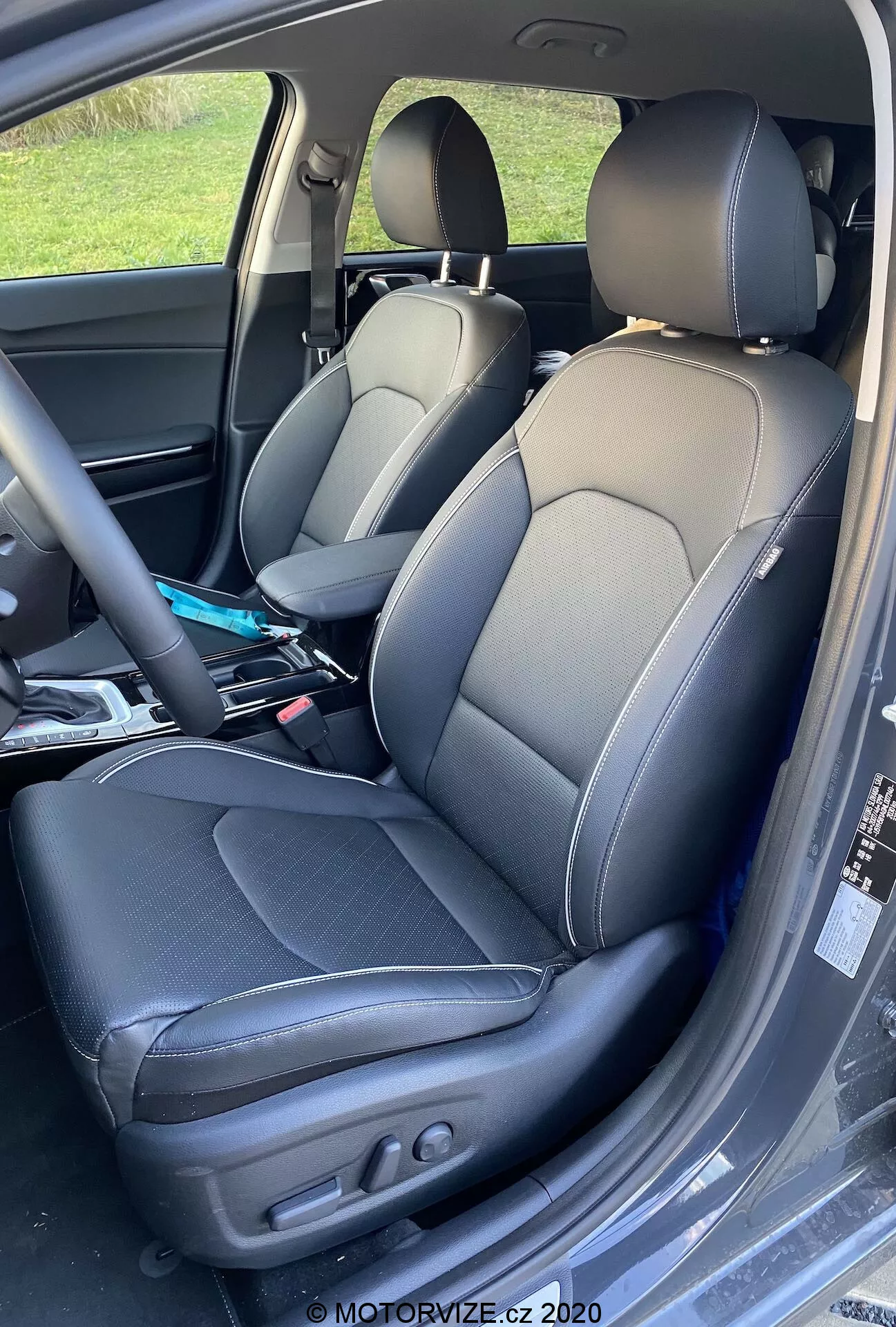 Detailný záber na interiér Kia Ceed 2018, zobrazuje vodičove a spolujazdcove sedadlá s dvojfarebným koženým čalúnením s kontrastným šitím, športovým dizajnom sedadiel s bočnými opierkami a elektrickým nastavením na vodičovom sedadle na ovládanie sklonu operadla a polohy sedadla. Jemné záhyby naznačujú používanie a na sedadle spolujazdca sú viditeľné rôzne predmety.